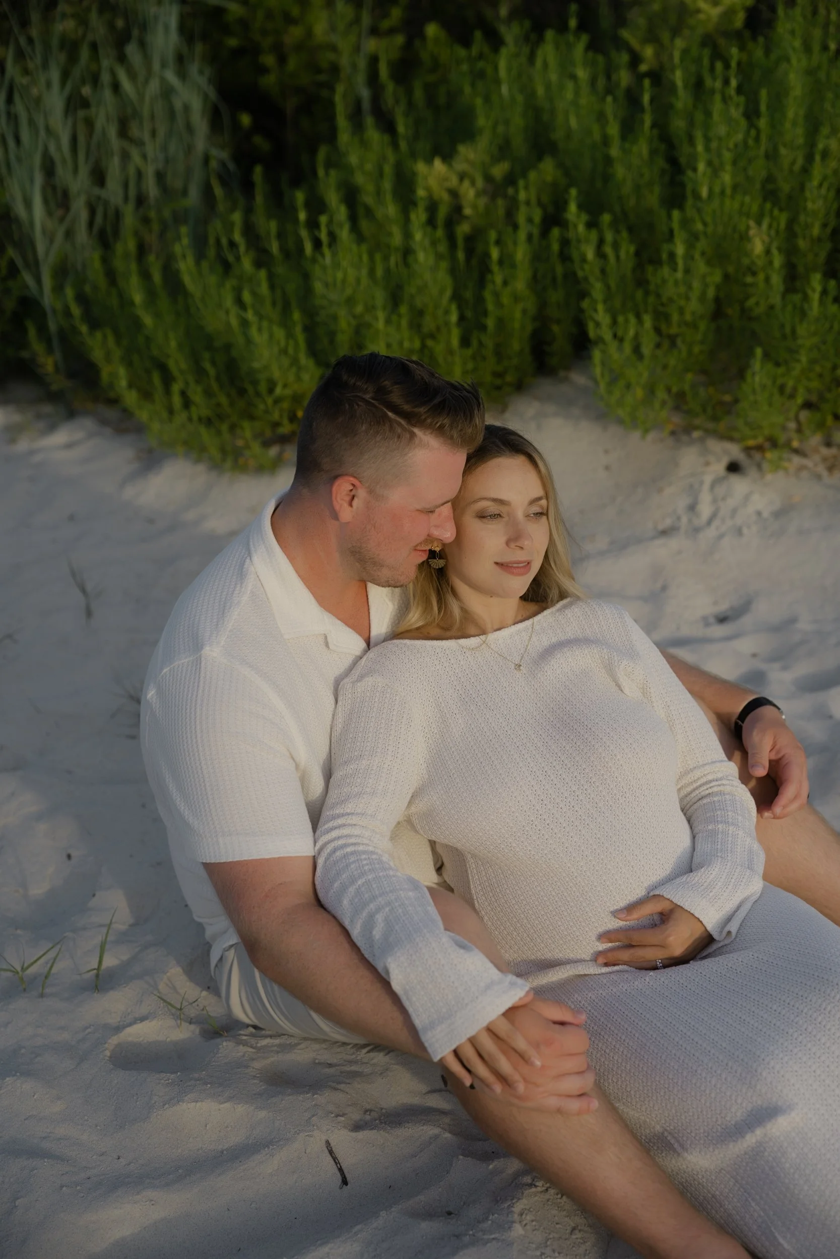 A couple sitting on the sand at the beach, with lush green plants in the background, during sunset