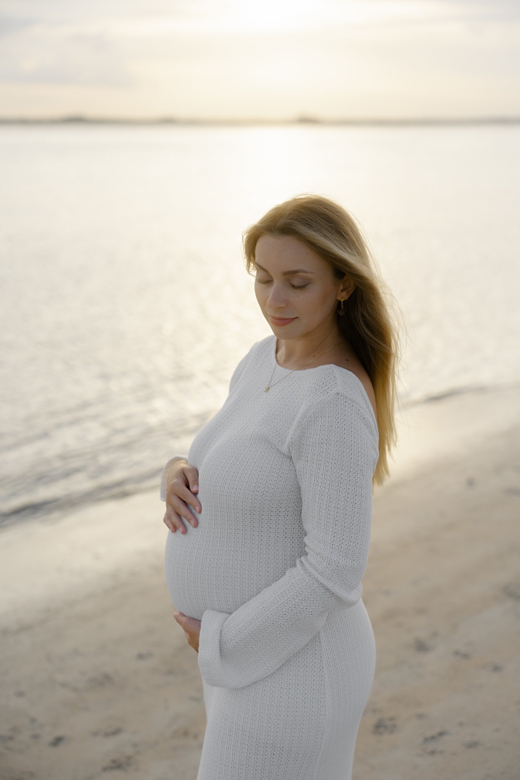 Pregnant woman in white dress stands on beach with water and sky in background during sunset, eyes closed and hands on belly.