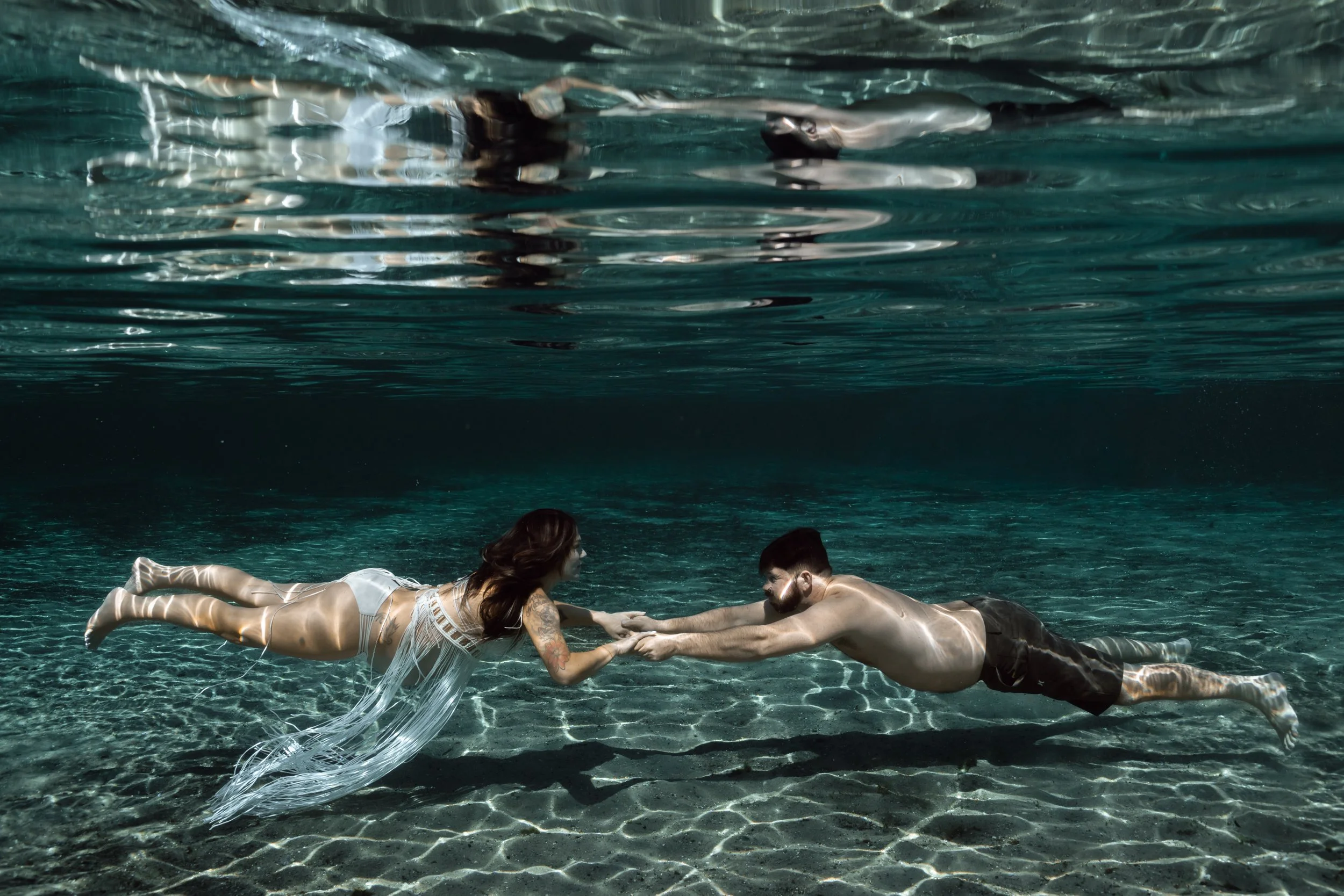 A man and woman holding hands underwater, swimming towards each other in a clear, dark aquatic environment.