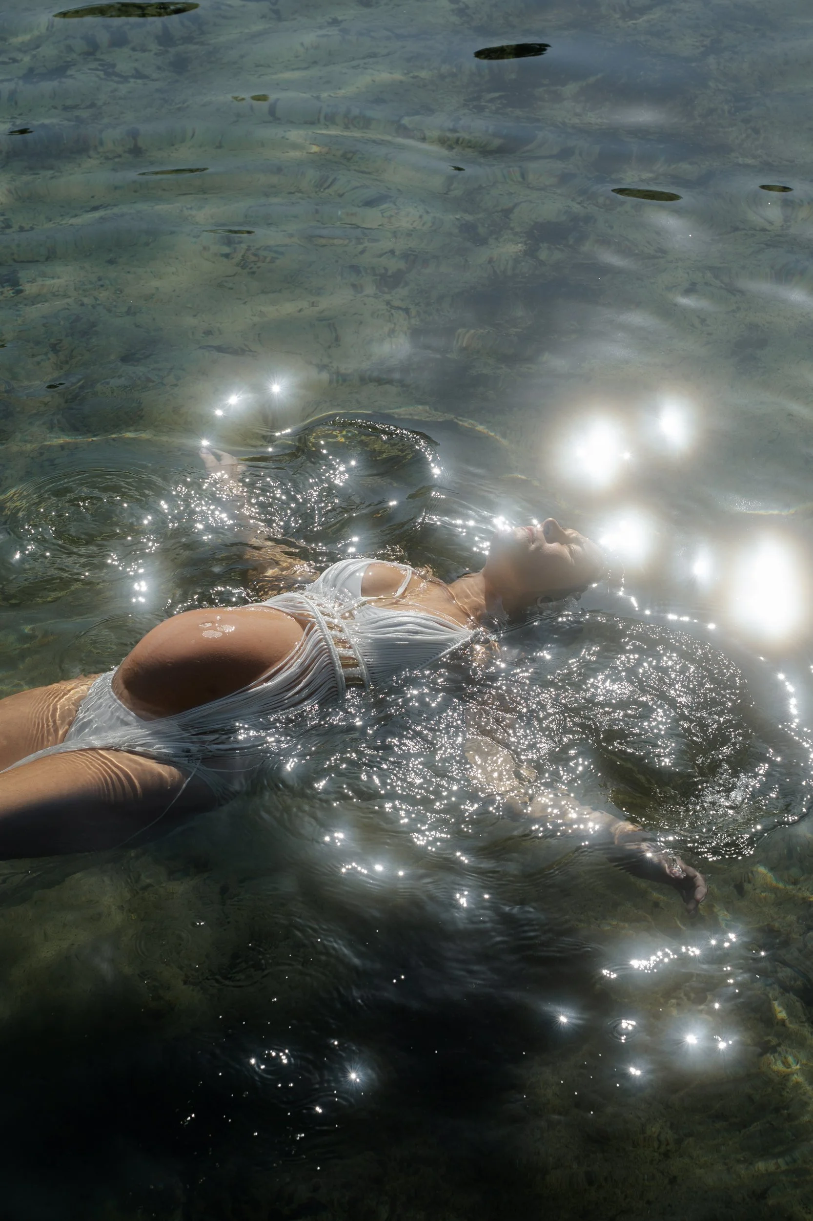 A woman in a white swimsuit floating on her back in clear water with sunlight reflections.