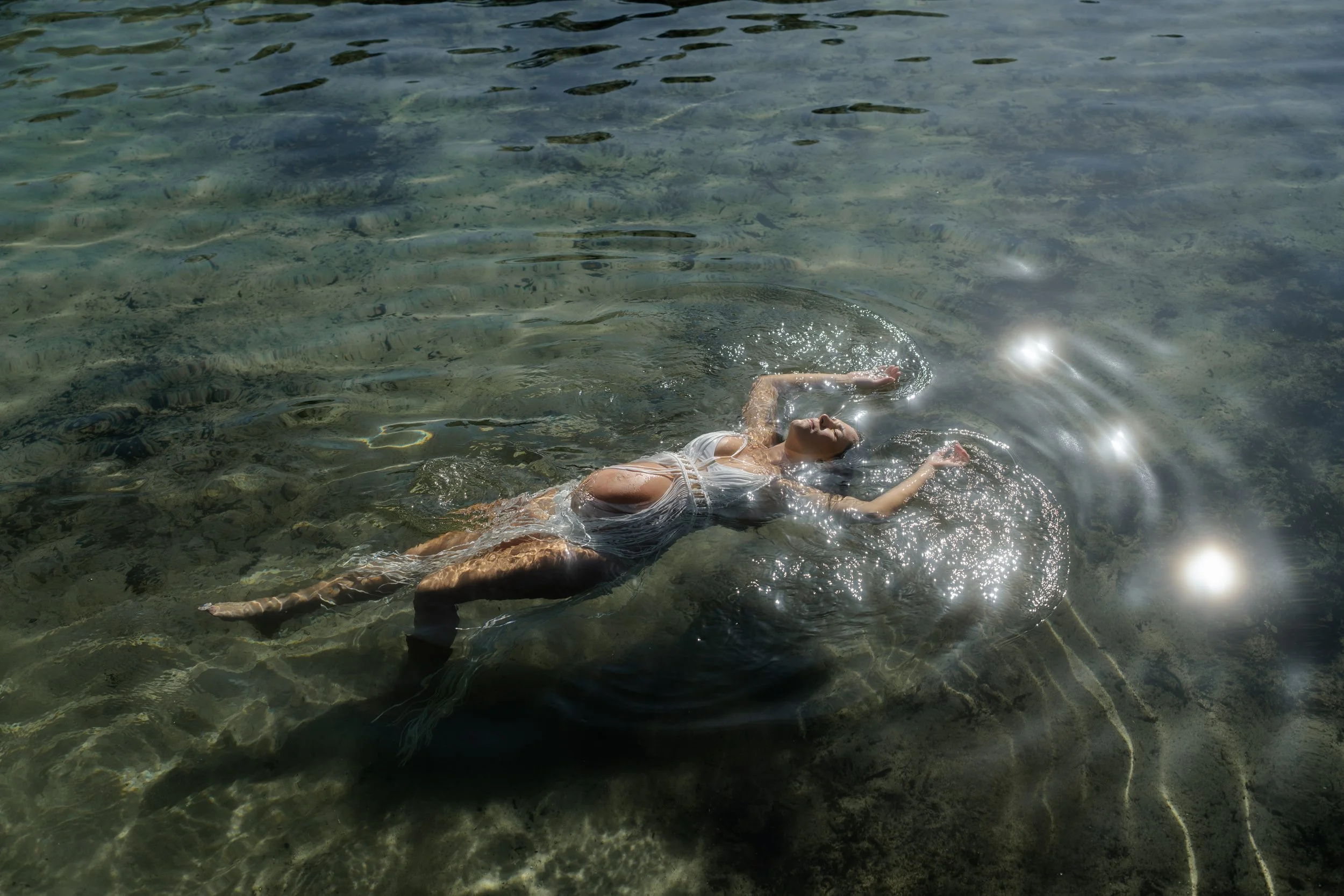 A woman floating on her back in clear water, wearing a white dress, with sunlight reflecting off the surface.