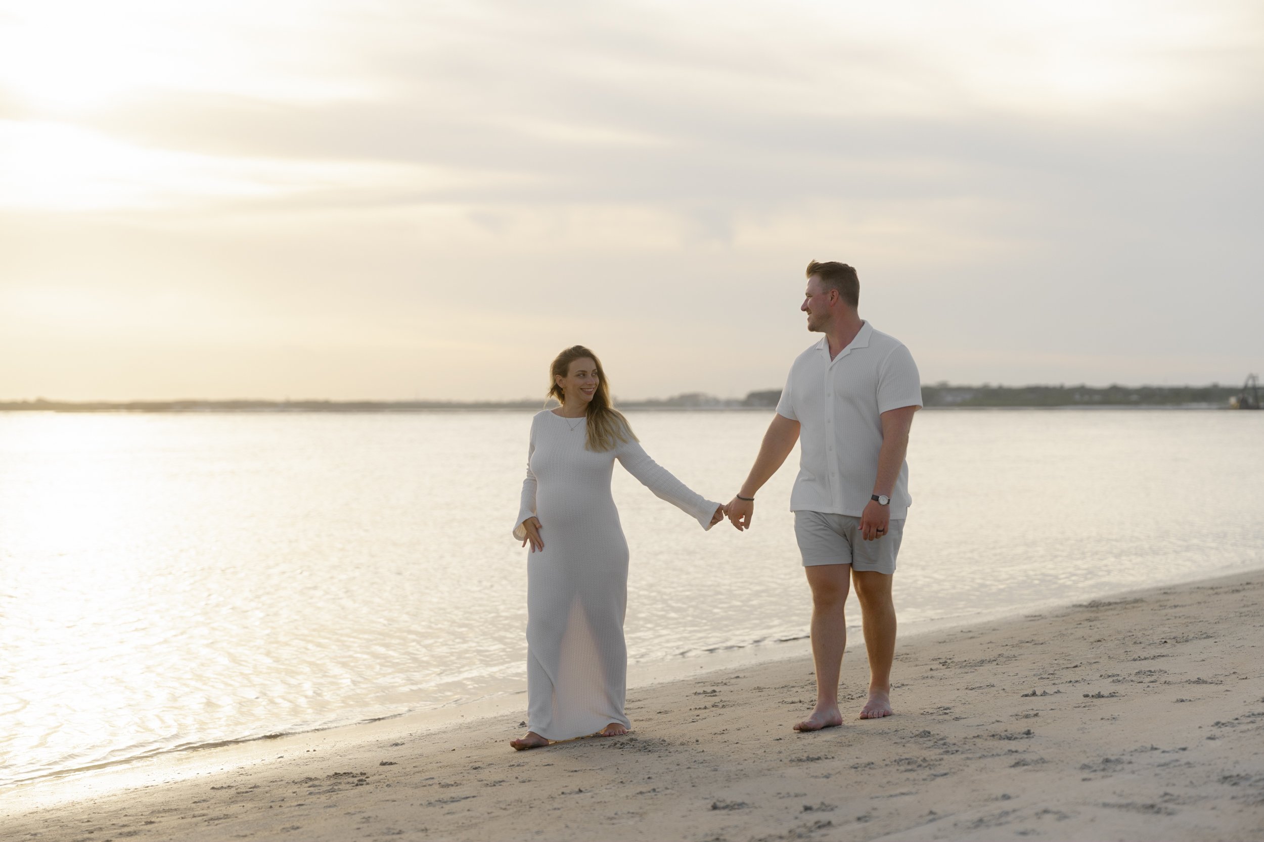 A couple holding hands and walking along a beach at sunset, with water and sky in the background.