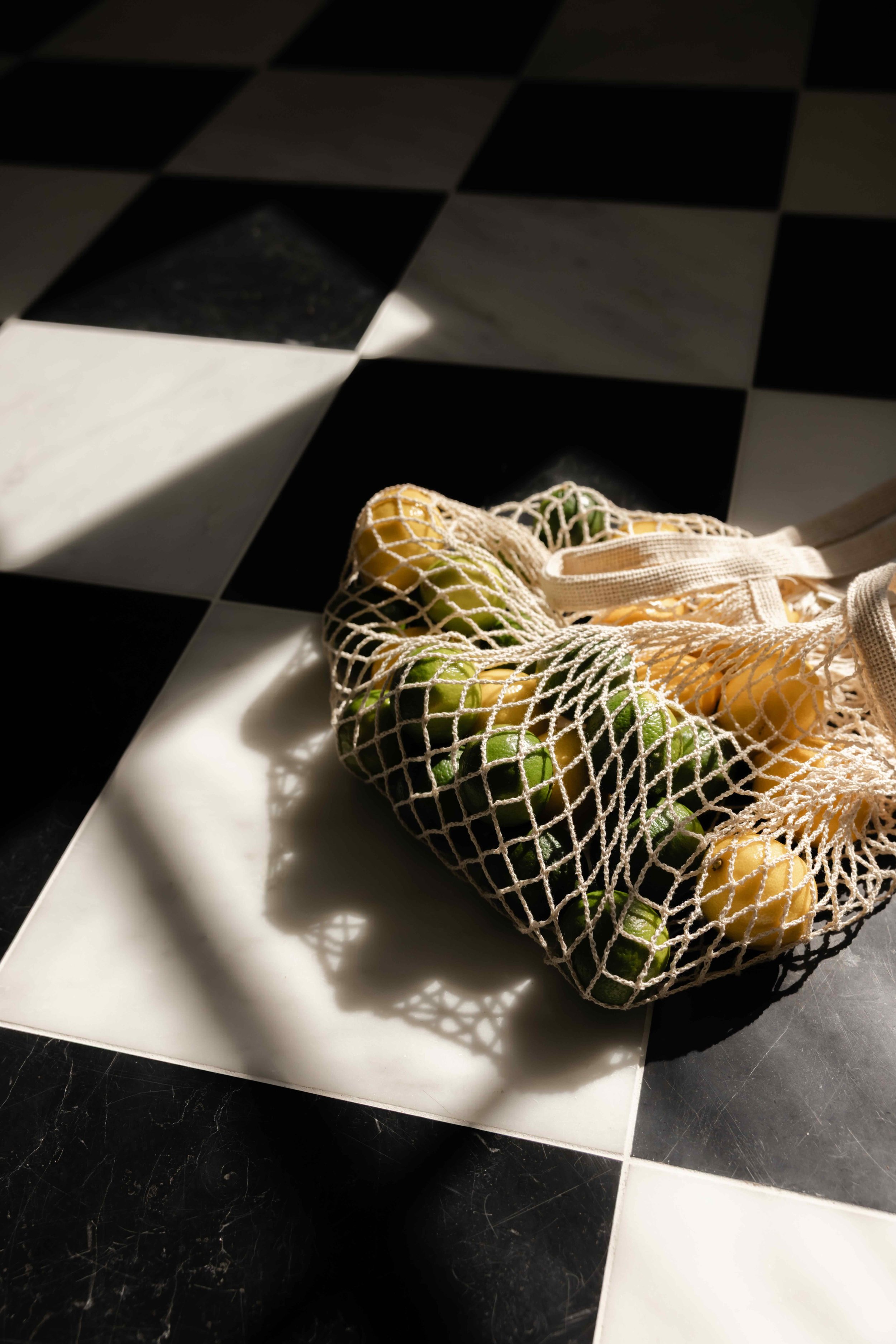 A reusable mesh bag containing yellow and green grapes lying on a black and white checkered floor, with sunlight casting a shadow from the bag.