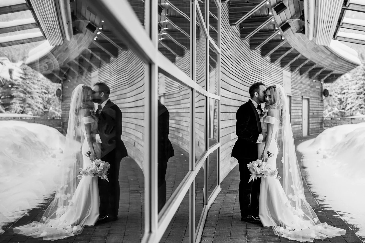 A black and white photo of a bride and groom sharing a kiss outside a modern building with glass windows and wooden siding, alongside a snow-covered path.
