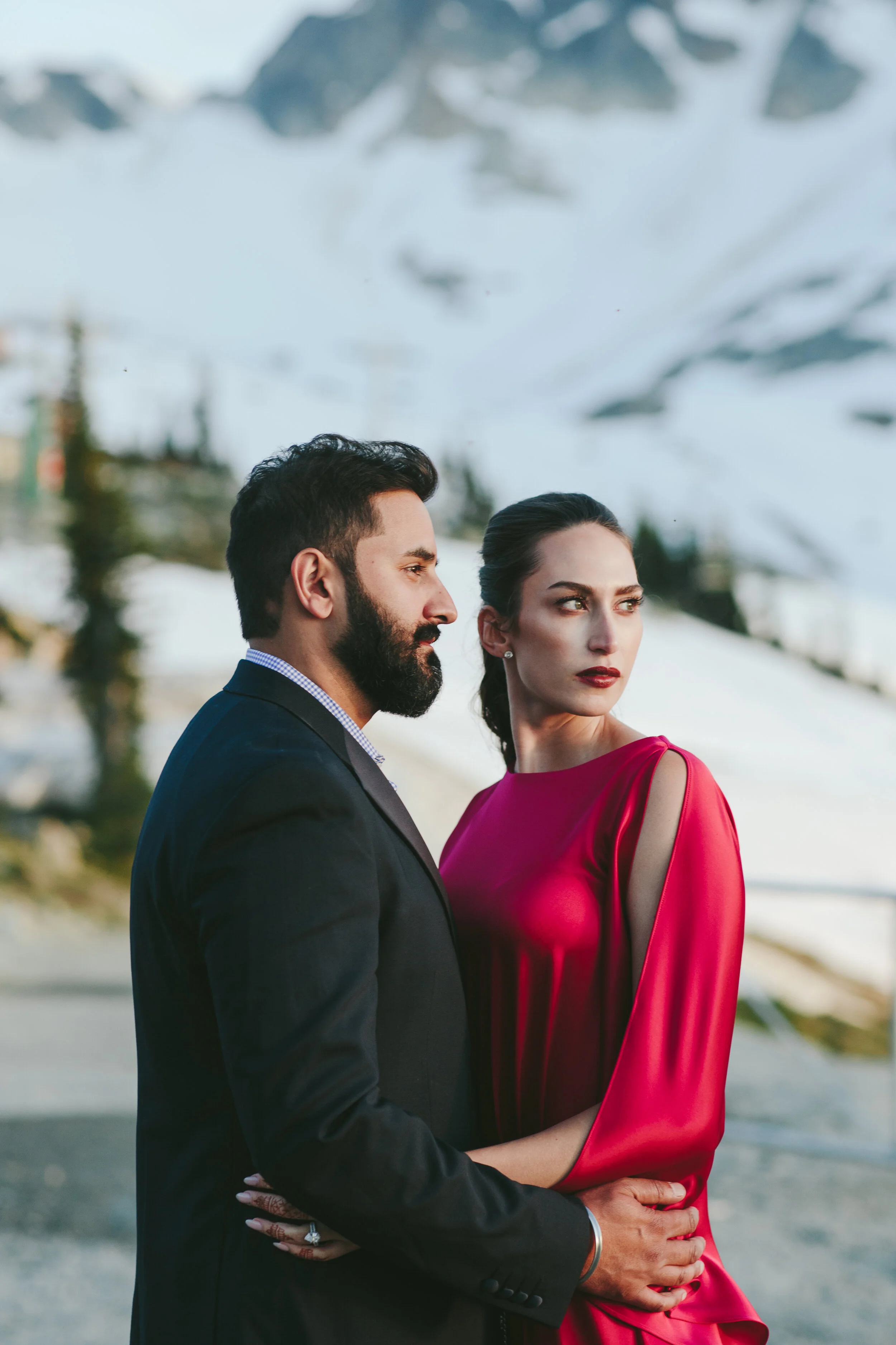A man in a suit and a woman in a red dress stand close together outdoors with snowy mountains in the background.