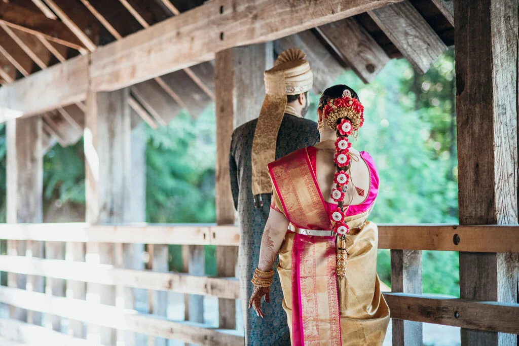 A bride and groom in traditional Indian wedding attire standing on a wooden bridge with a natural green background.