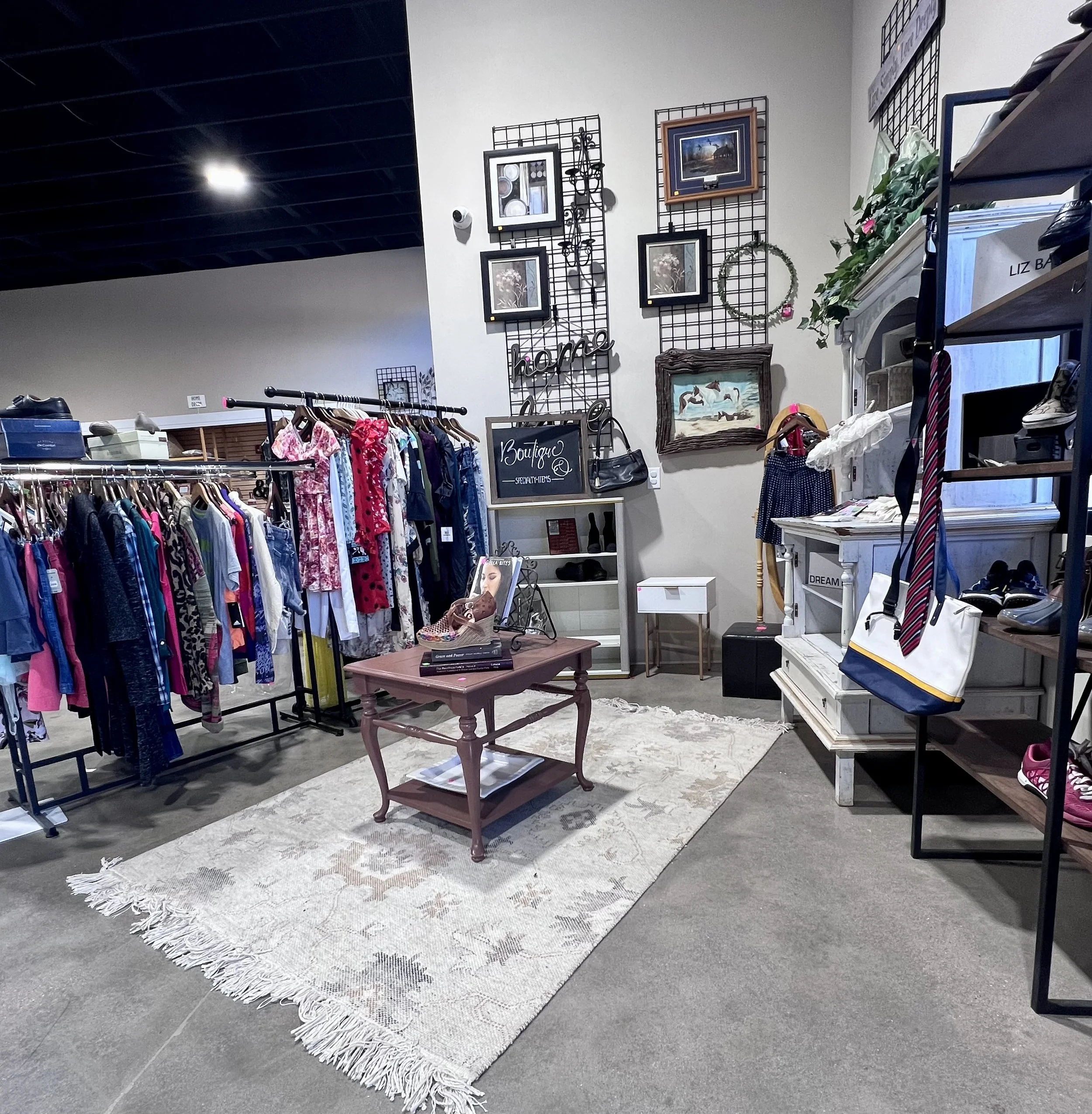 Interior view of a boutique or thrift store displaying clothing on racks, with artwork and decorative items on the walls, a vintage-style rug on the floor, and a shelving unit with shoes and boxes.