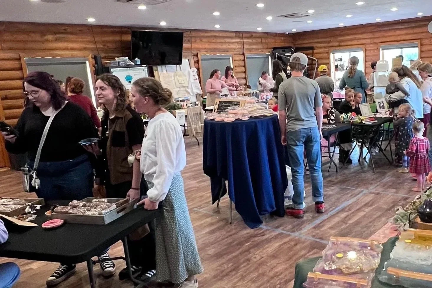 A craft fair or market inside a wooden cabin with people browsing tables of handmade goods and baked items, and children with adults browsing and shopping.