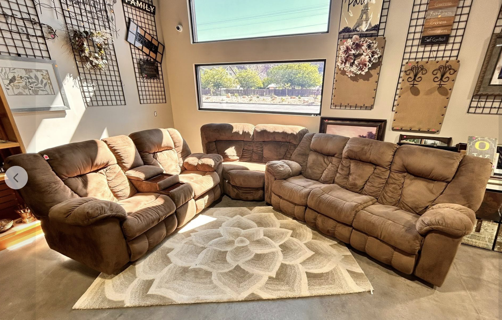 A cozy living room with large brown sectional sofas arranged in a U-shape, a patterned area rug on the floor, and two large windows allowing natural light. The walls are decorated with various wall art, including floral and Eiffel Tower themed pieces, and decorative metal wall hangings. Outside the windows, green trees and a backyard are visible.