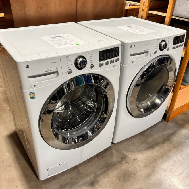 Two front-loading washing machines, one LG and one another brand, placed side by side in a store or showroom with wooden shelves nearby.