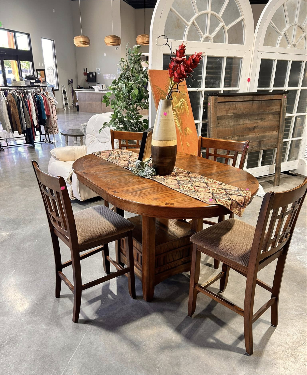 A wooden dining table with four chairs, a decorative runner, a tall vase with red flowers, and a framed picture. In the background, there are retail clothing racks, a couch, plants, and a counter with hanging pendant lights.
