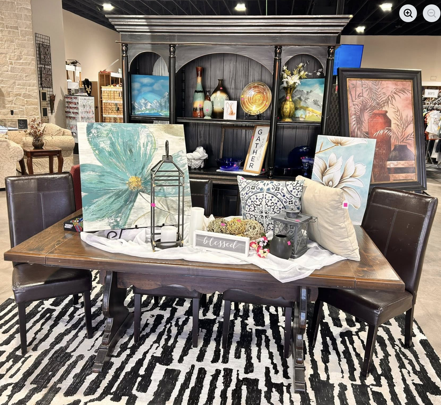 A display table in a store with various home decor and artwork, including paintings of flowers and landscapes, decorative vases, a lantern, a sign reading 'blessed', and pillows, all arranged on a black and white patterned rug.