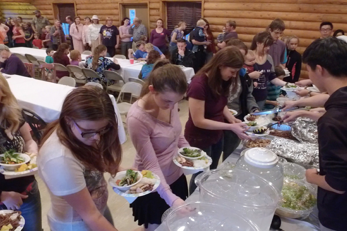 People in line at a buffet table with food, in a room with wooden log cabin walls, some seated at tables and others waiting in line for food.
