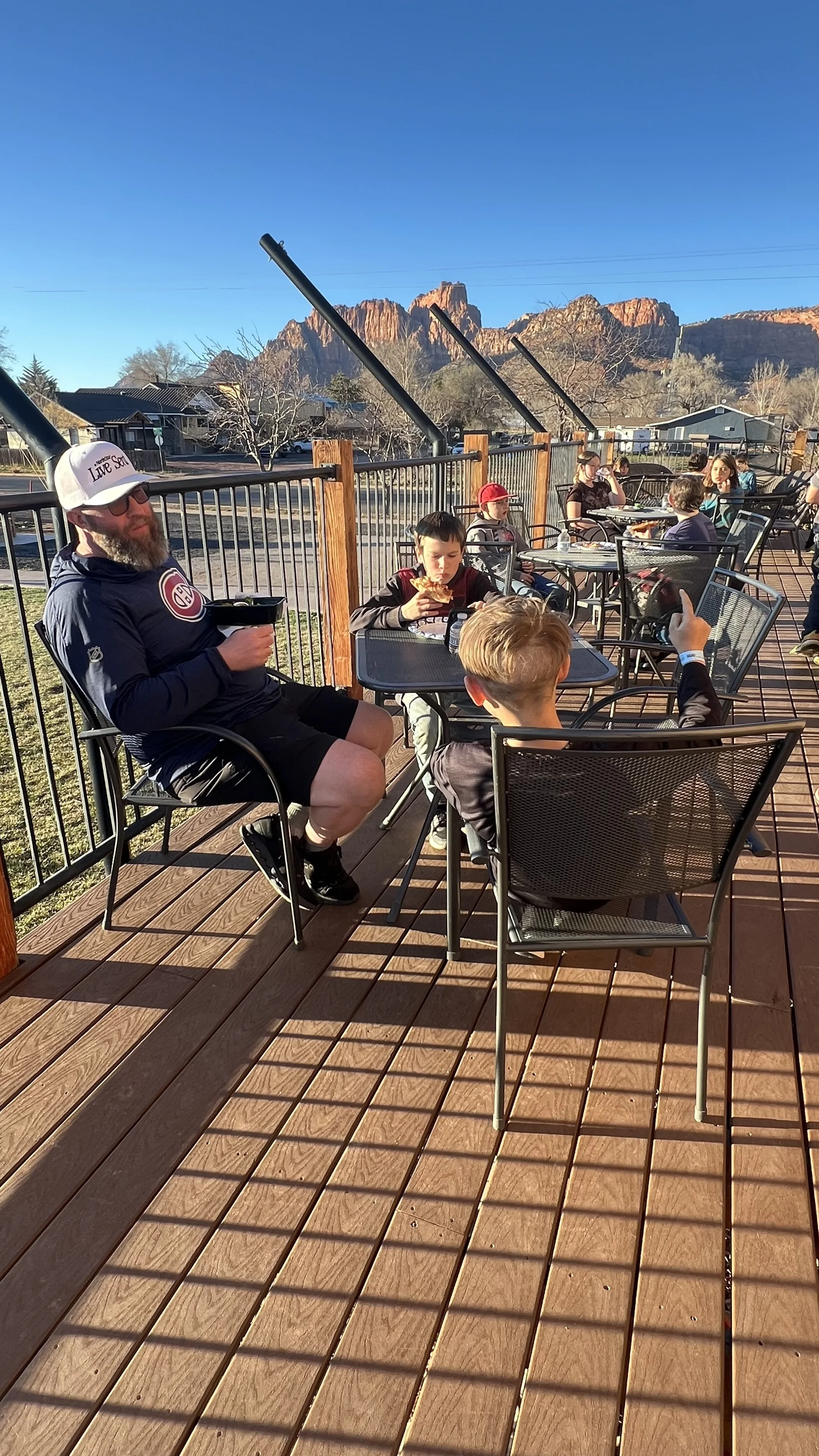 People sitting on a wooden outdoor patio with mountains in the background, some eating, talking, and pointing.