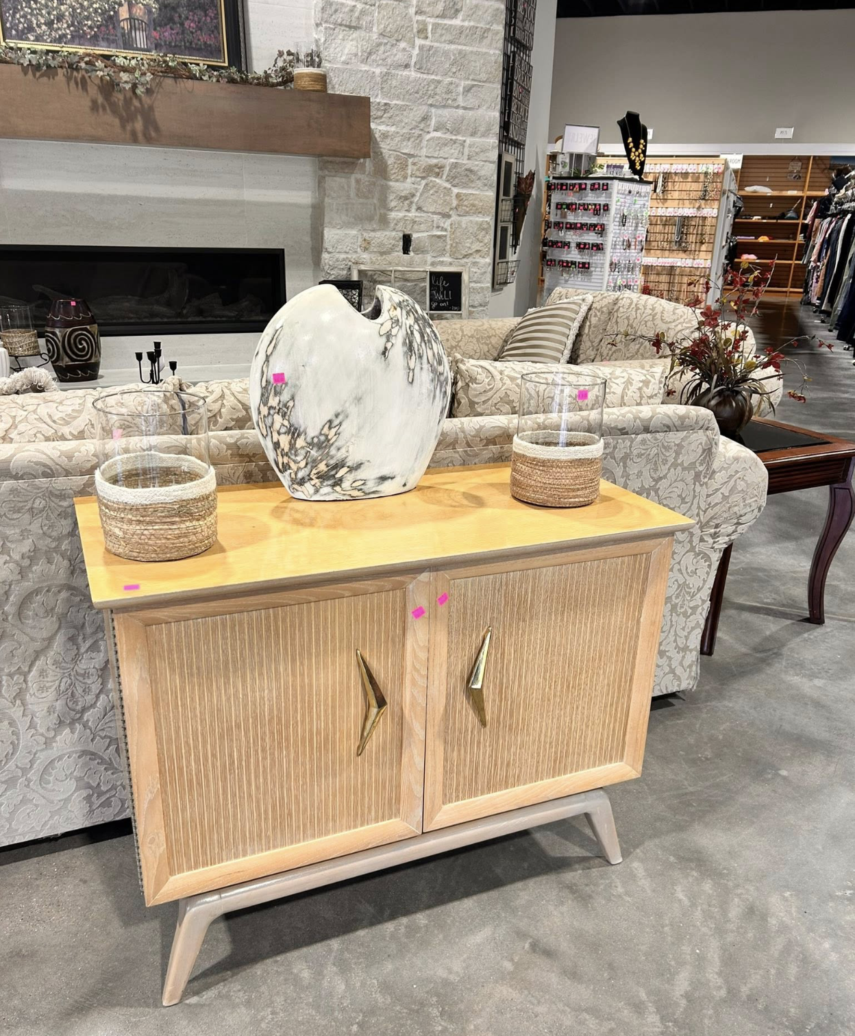 A wooden sideboard with golden handles displaying a large abstract ceramic sculpture and two glass vases with woven bases. Behind, a beige patterned sofa with striped and floral pillows is visible, along with a decorative potted plant and a fireplace with a white stone surround. In the background, a retail store with jewelry, clothing, and accessories on display can be seen.