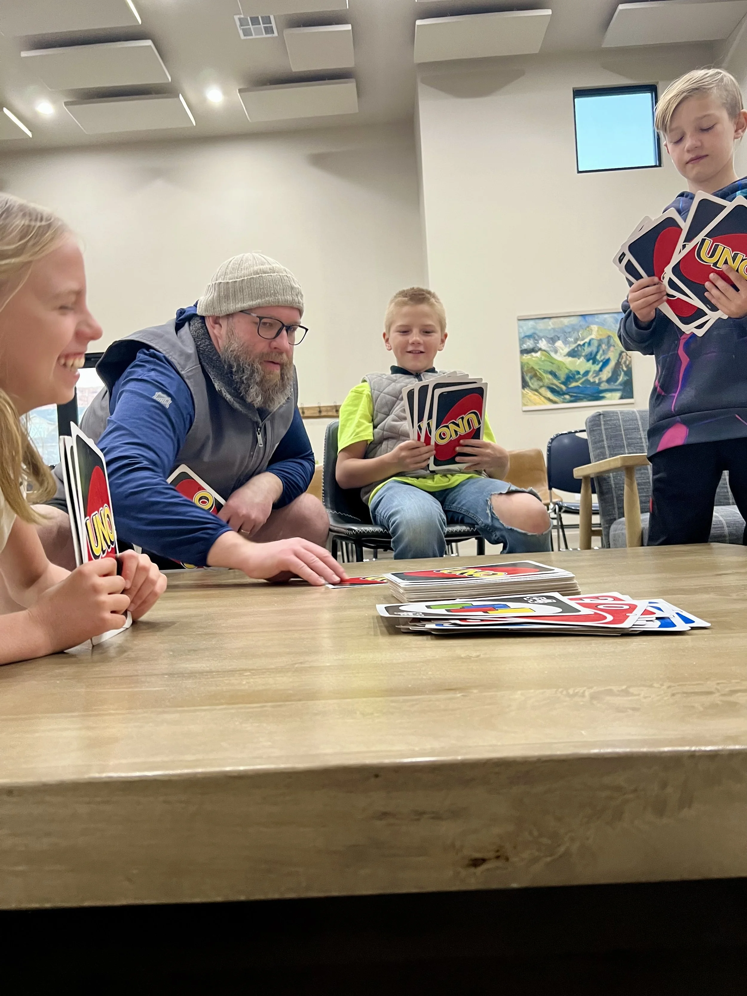 Four people sitting around a wooden table playing the card game UNO, with a stack of UNO cards on the table, inside a room with a painting on the wall and ceiling lights.