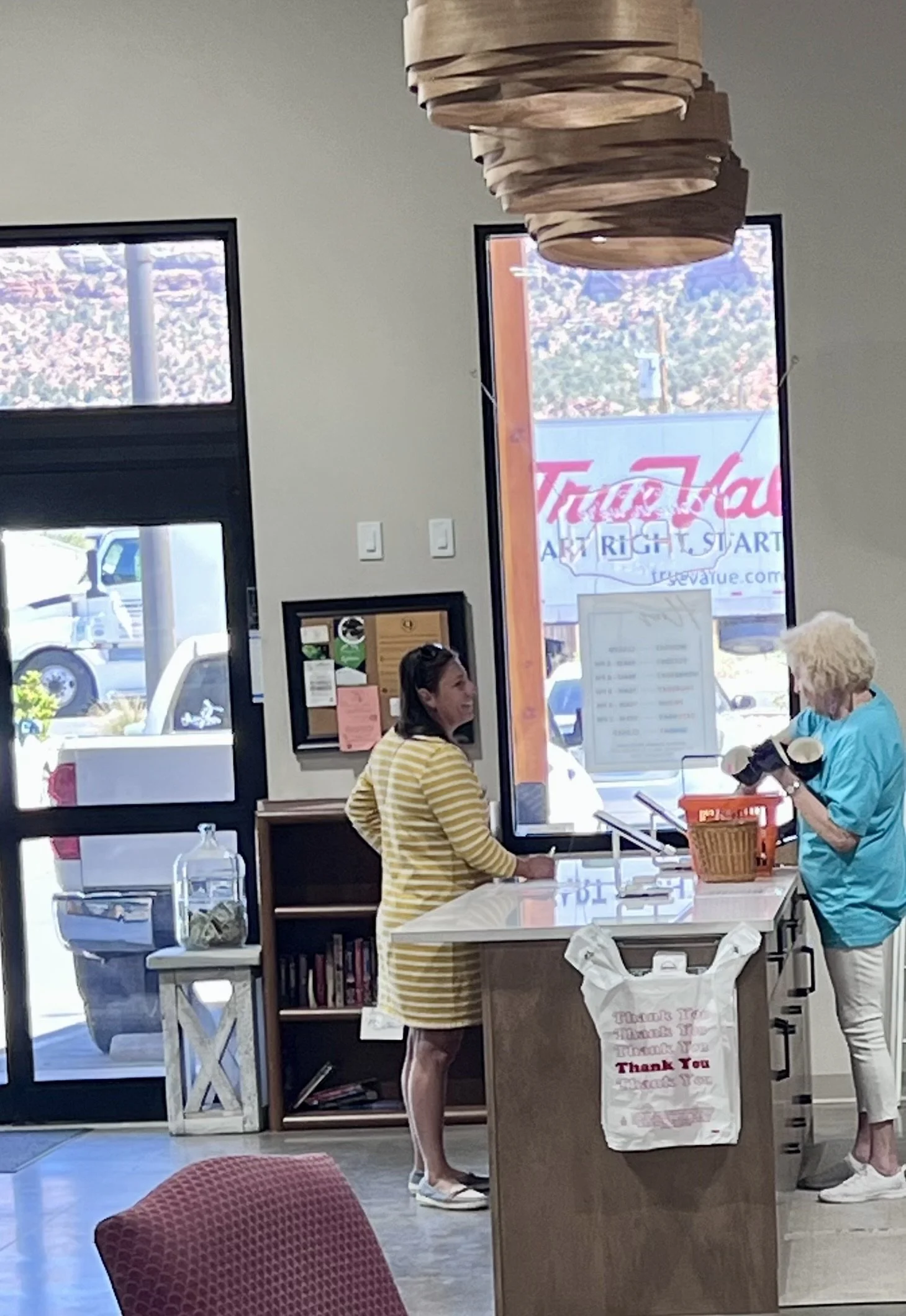 Inside a store with two women at the counter, one in a yellow striped dress and the other in turquoise, who is holding a camera. Large windows with signage outside, a red chair in the foreground, and various items on the counter and shelves.