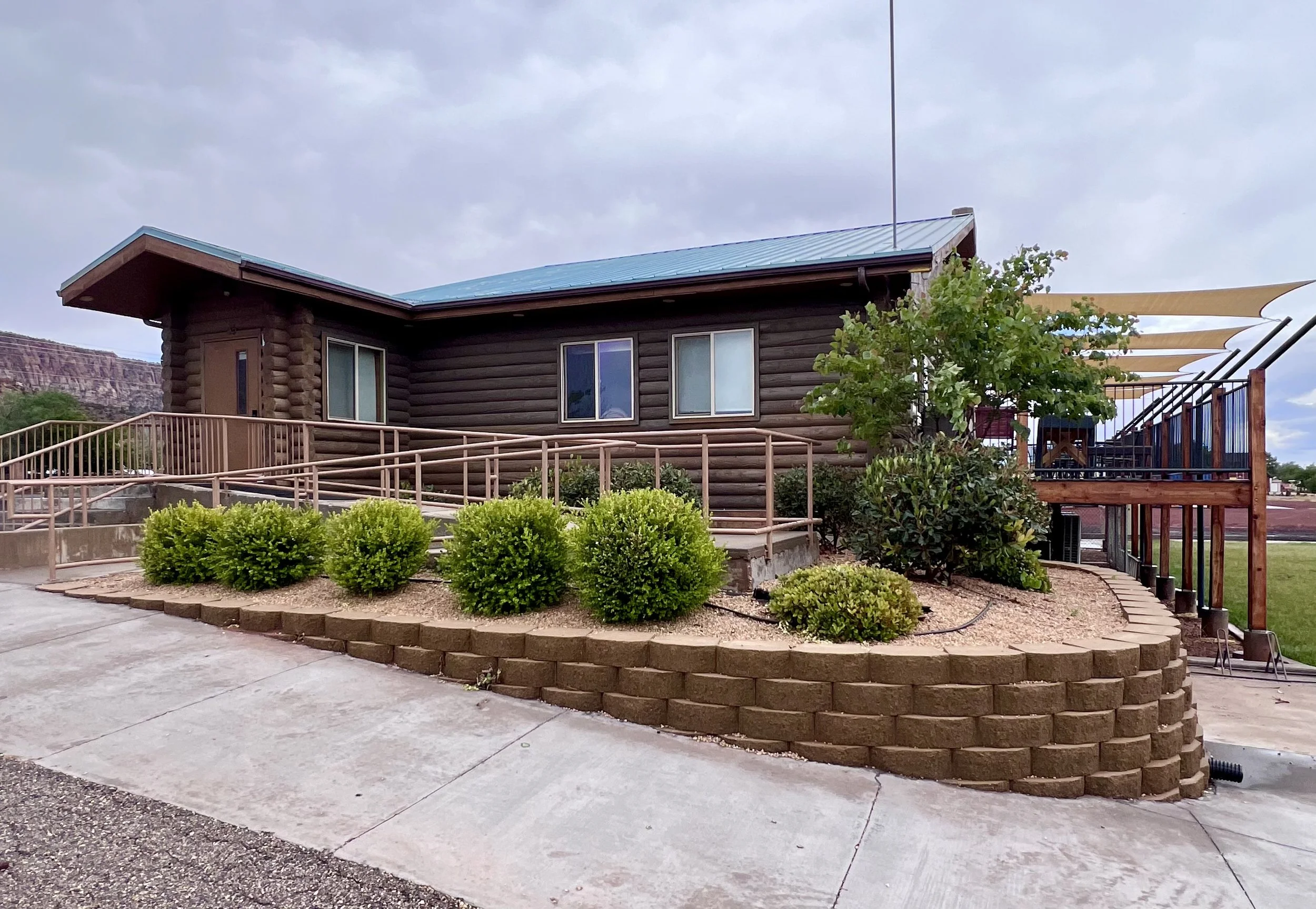 A brown log cabin-style building with multiple windows and a metal roof. The building is surrounded by landscaped bushes, a curved brick retaining wall, and a concrete patio with a ramp and steps. There is a tree and some shade sails on a deck to the right of the building, with mountains visible in the background under a cloudy sky.