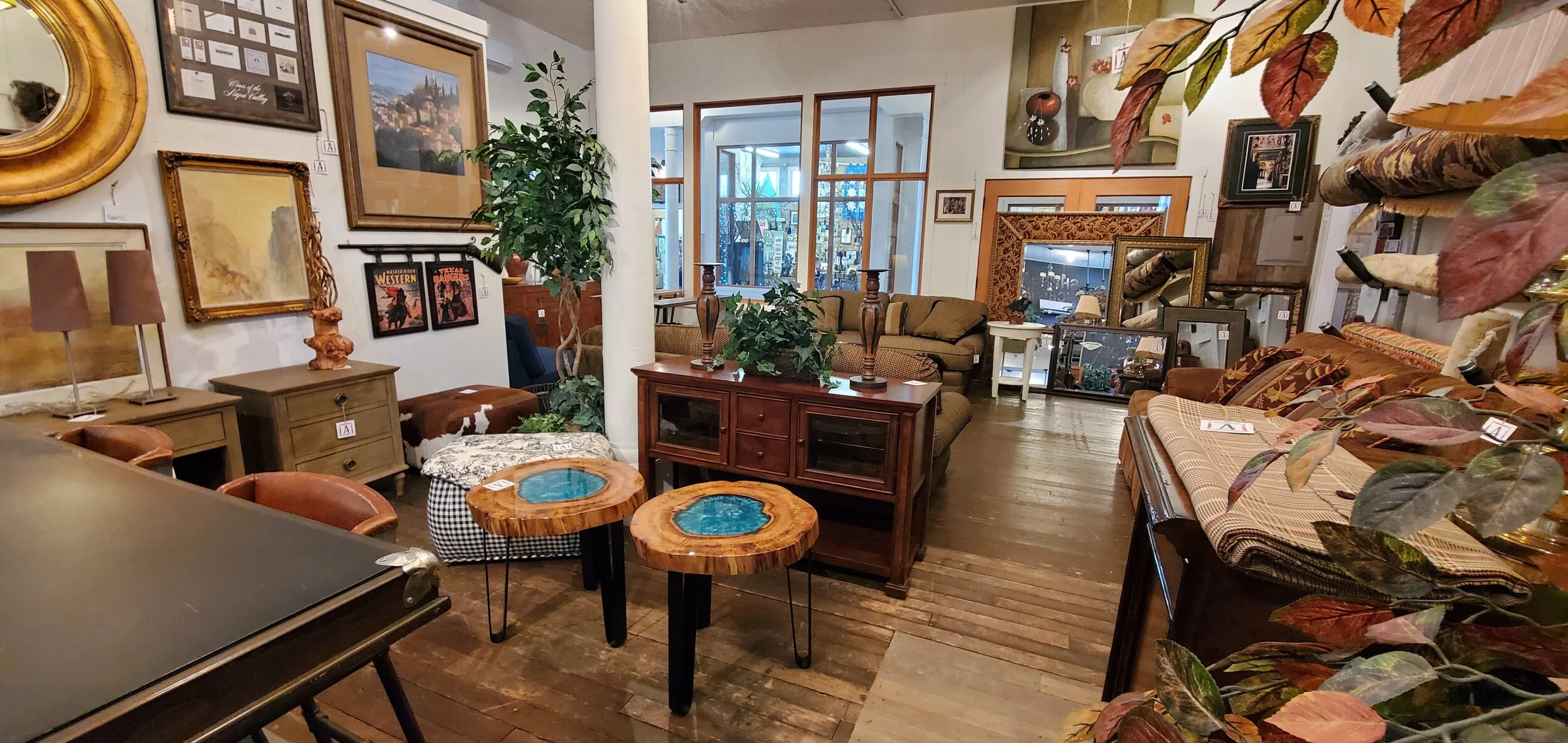Interior of an art and furniture store with various paintings on the wall, wooden furniture, a sofa, plants, and decorative items.