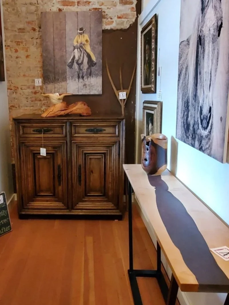 An art gallery corner with various paintings and sculptures, including a horse riding figure on a wooden panel, a bird sculpture, a wooden mask, and framed artwork on the wall, along with a wooden cabinet and a table.