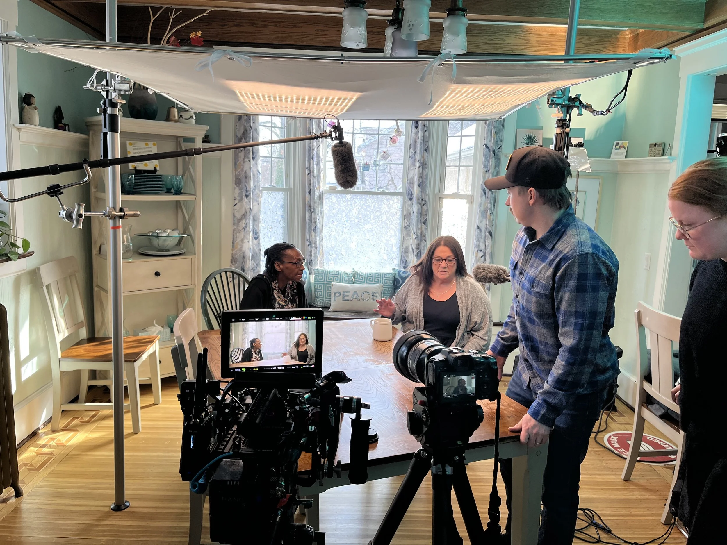 A behind-the-scenes photo of a video production, showing two women and two crew members in a brightly lit dining room, with a camera on a tripod and boom microphone overhead.