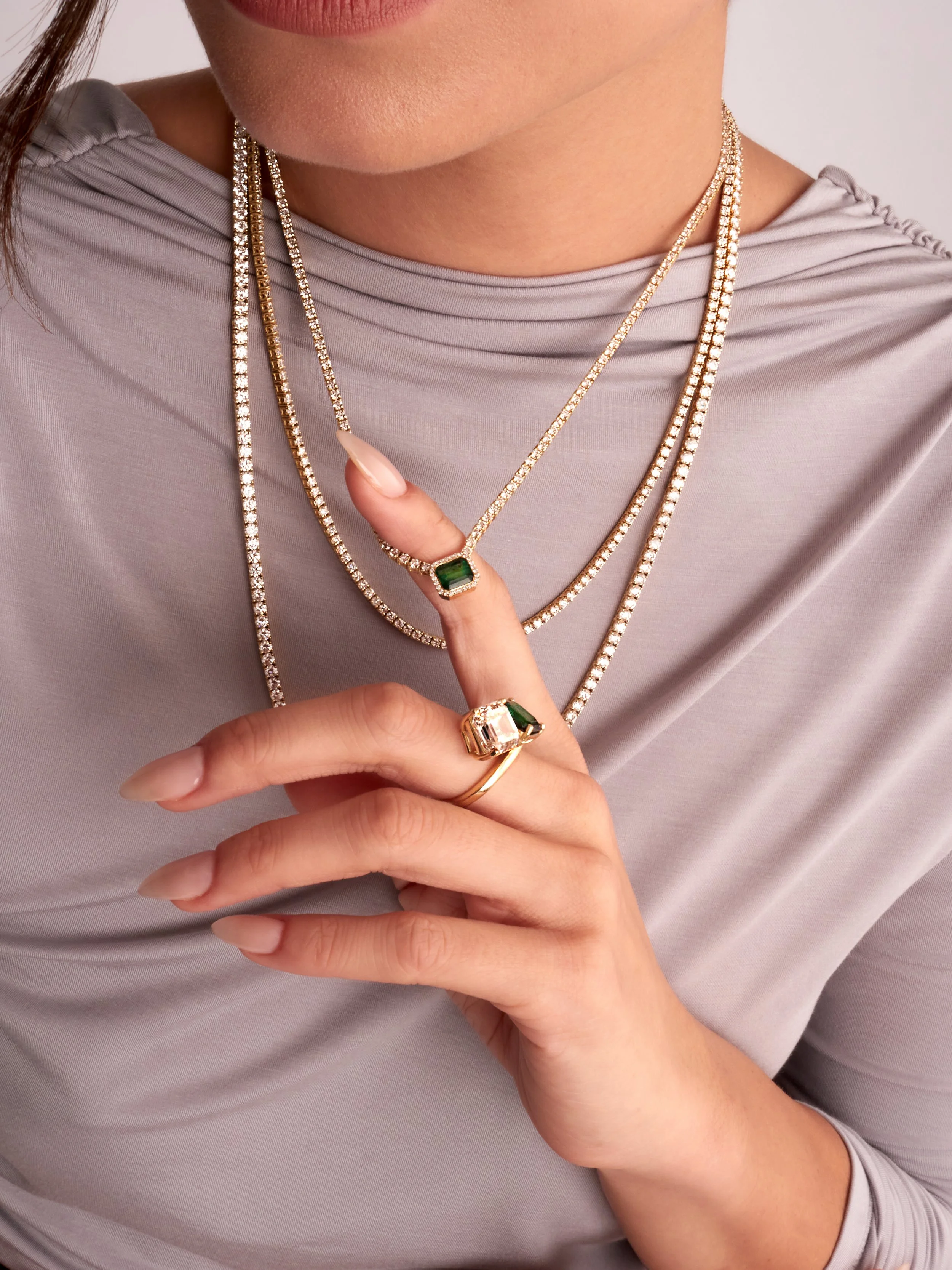 Close-up of a woman wearing layered diamond necklaces and rings with emerald and white stones, against a light gray top.