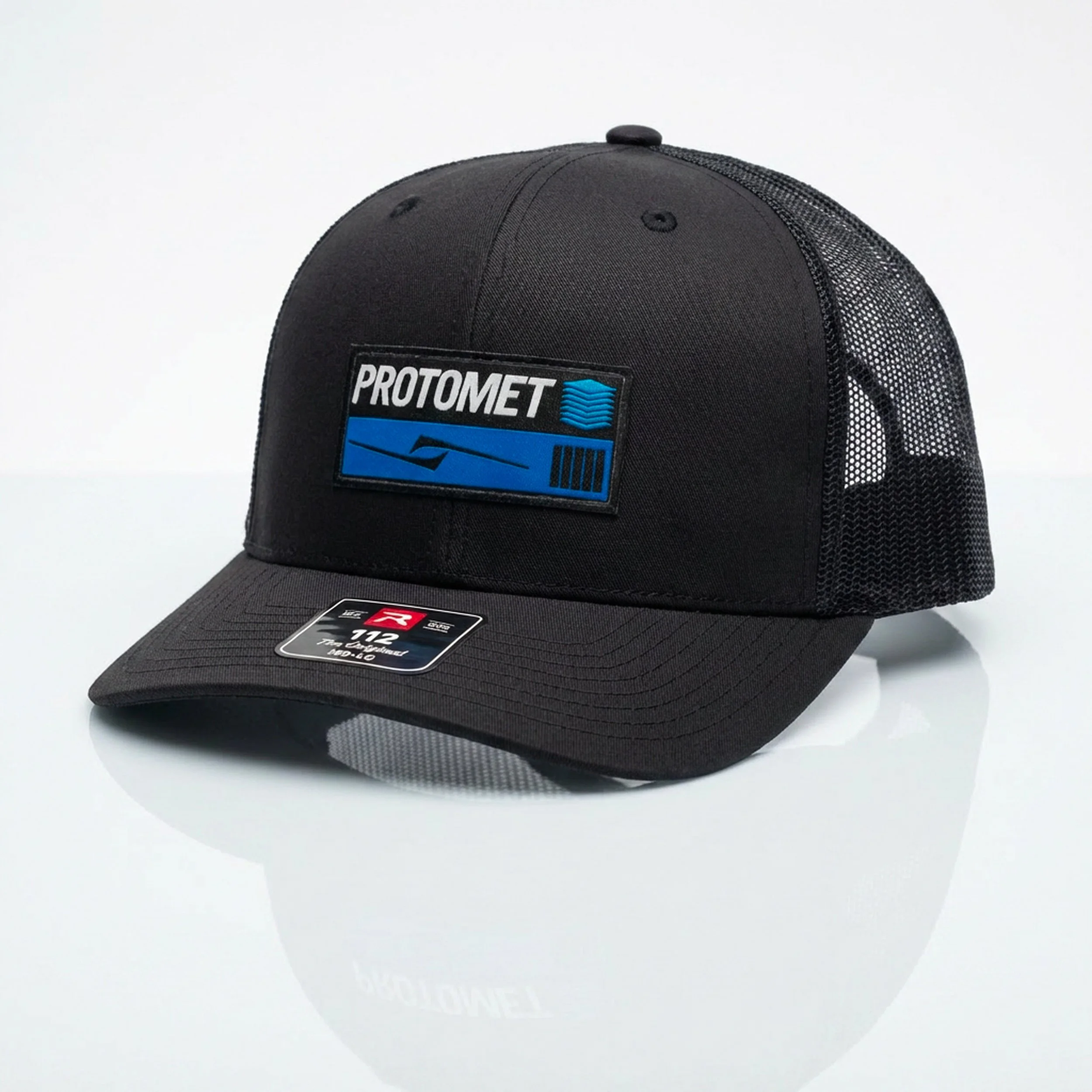 Black trucker hat with a mesh back, featuring a ProtoMet logo patch on the front panel, placed on a plain white surface.