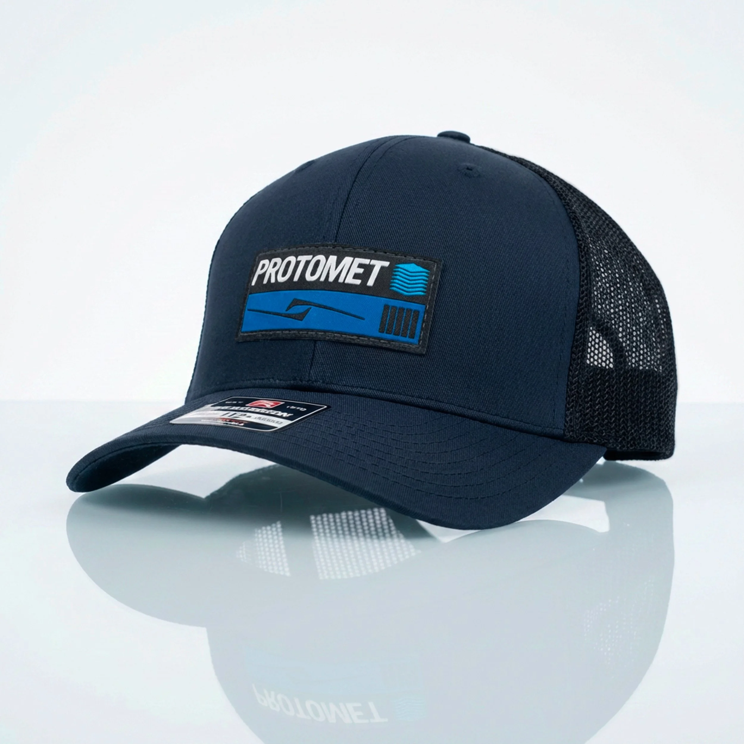 Close-up of a dark blue baseball cap with a "Protomete" logo, placed on a reflective surface against a plain white background.