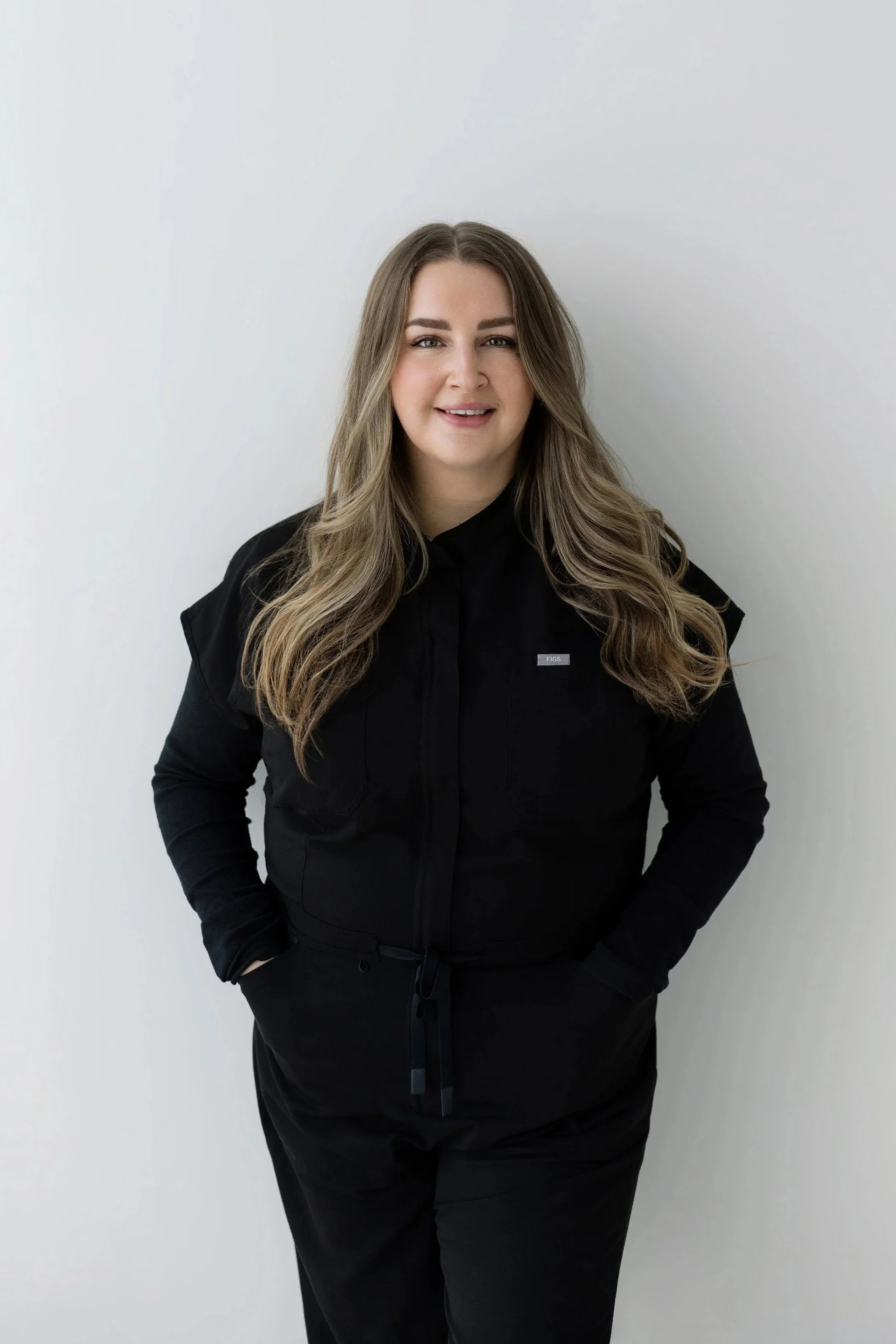 A young woman with long blonde hair, smiling and winking, wearing a black medical uniform with tattoos on her arms, standing against a plain white wall.
