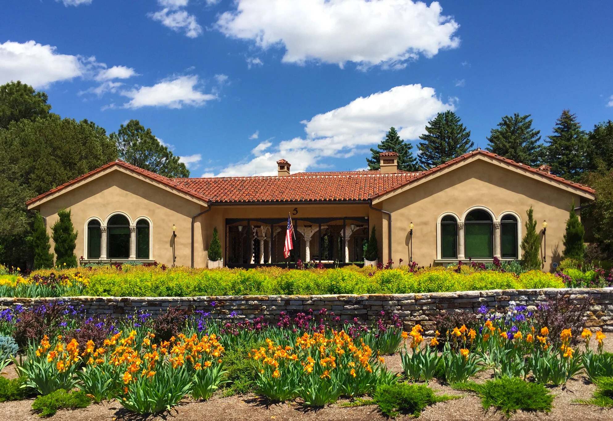 A Mediterranean-style villa custom designed by LGA Studios in Colorado Springs with adobe walls, a terracotta roof, and arched windows, surrounded by vibrant gardens with yellow and purple irises.