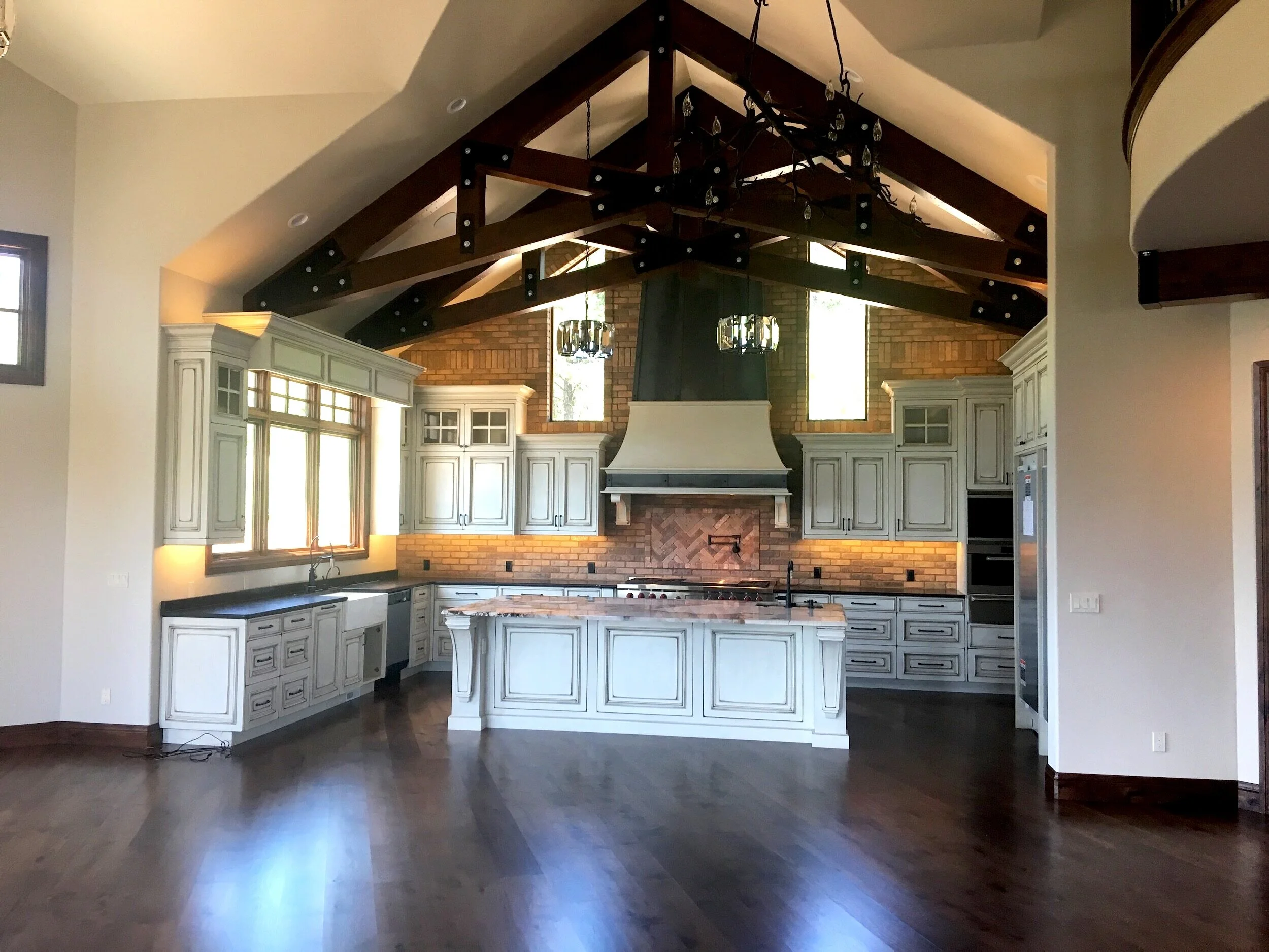 Spacious kitchen with vaulted ceiling, wooden beams, white cabinets, island with countertop, brick backsplash, large windows, and hardwood floors.
