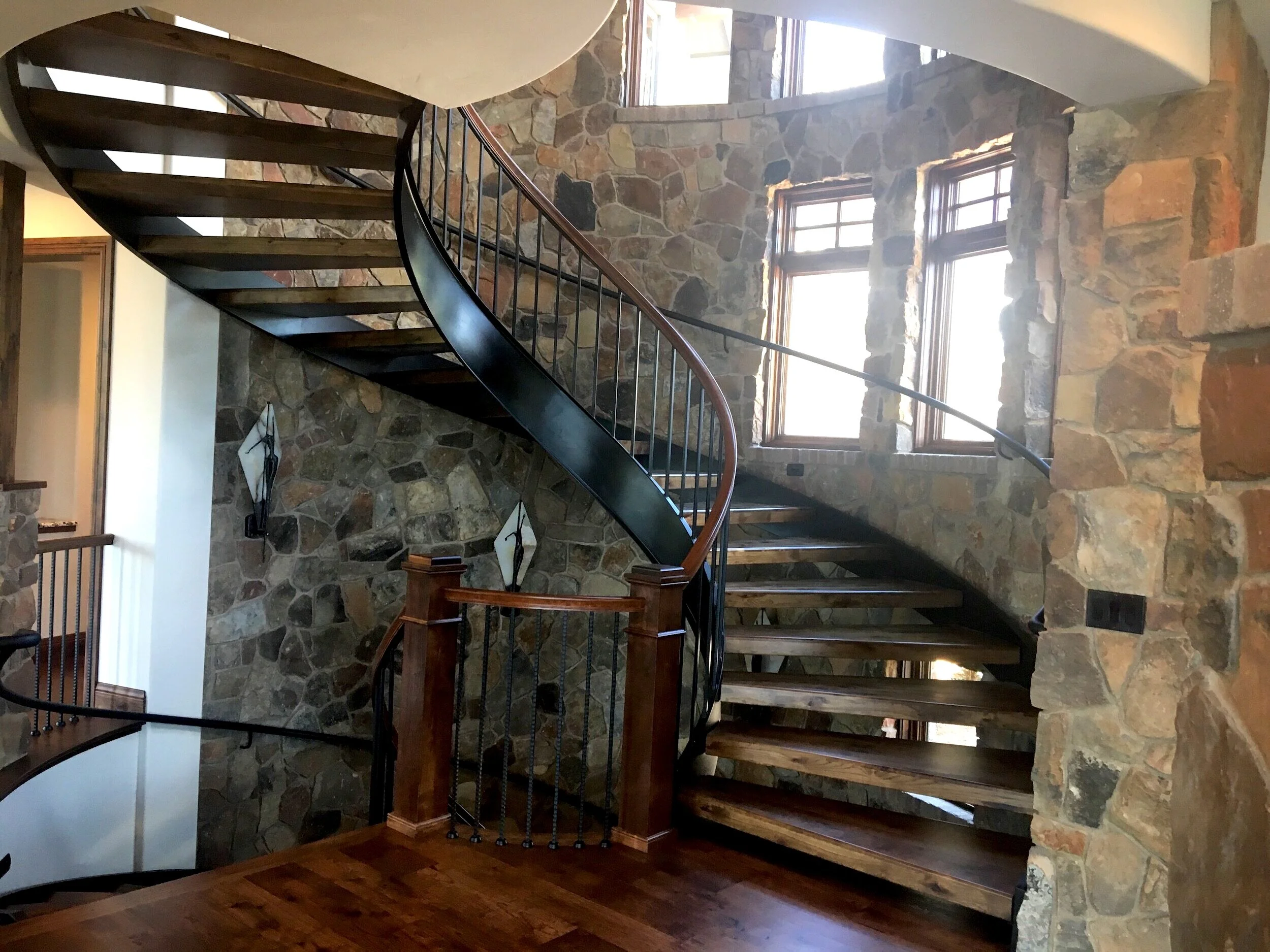Luxury custom home designed by LGA Studios with a spiral staircase with dark wood steps and metal railing, surrounded by stone walls and large windows in a Tuscan style. 
