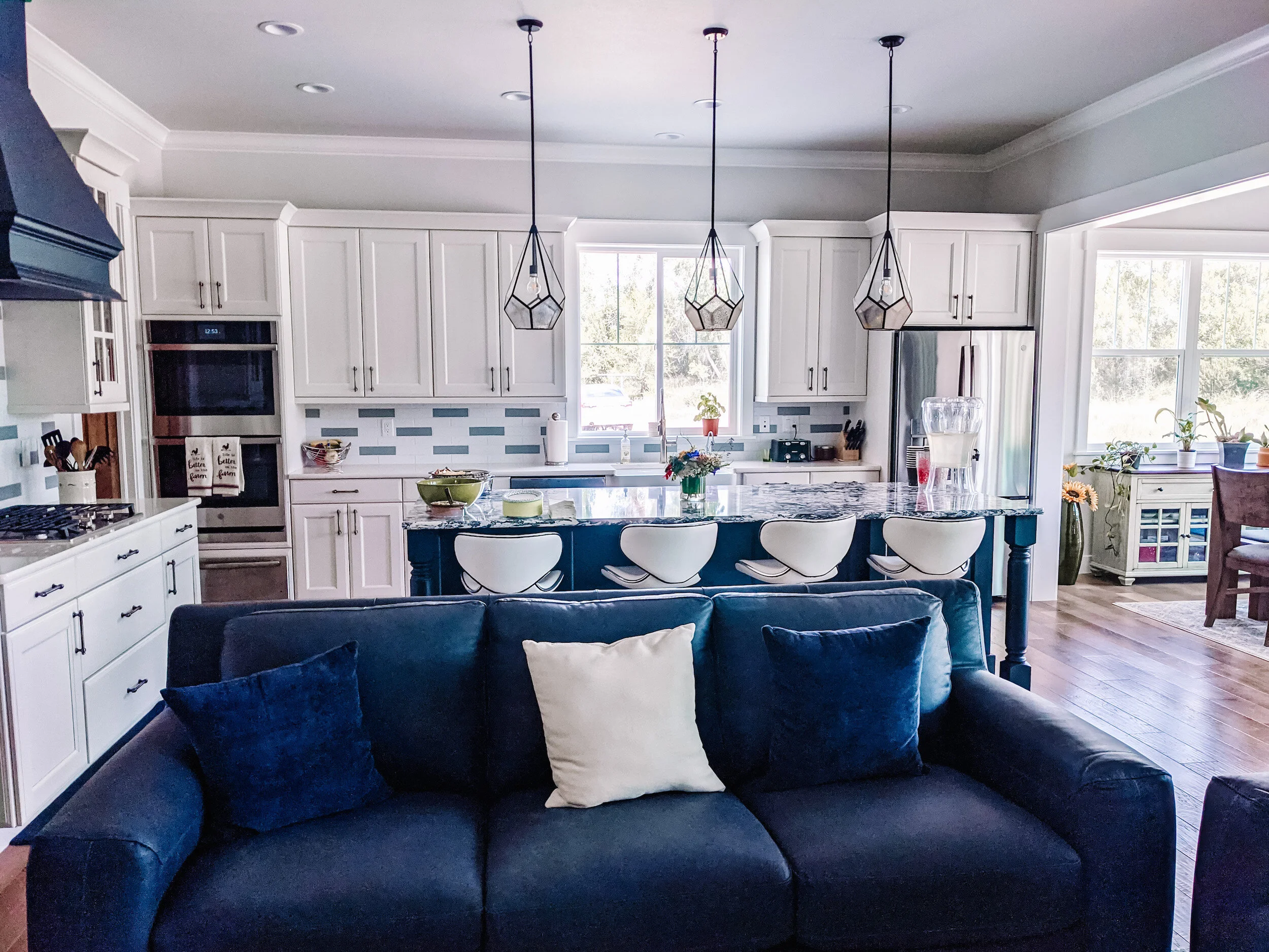 Modern farmhouse kitchen with white cabinets, island with granite countertop, pendant lights, blue velvet sofa, and blue pillows. Custom home design by LGA Studios.
