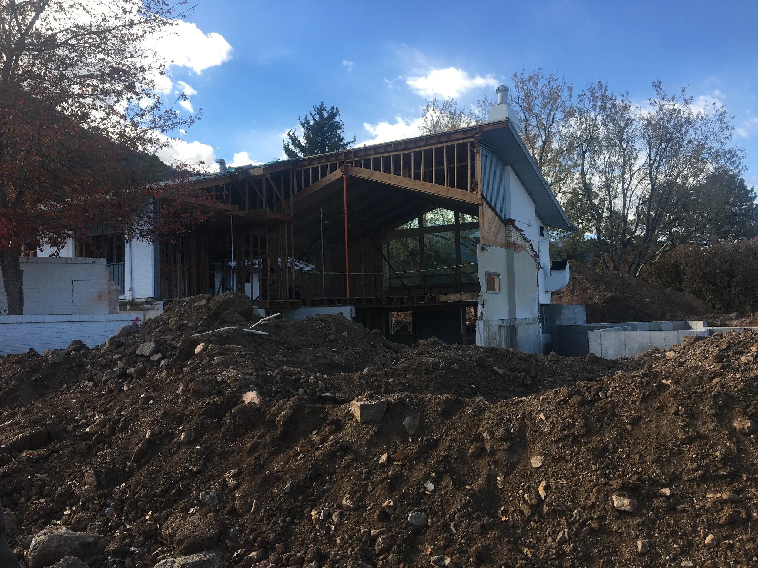 Partially constructed building site in Colorado with exposed wooden framework and mounds of dirt, set against a clear blue sky background with trees.