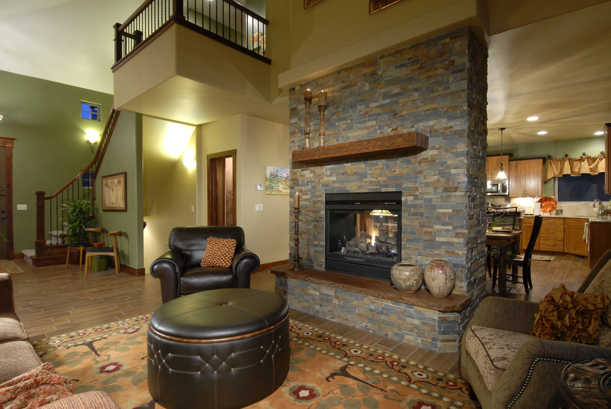 Colorado Springs Parade of Homes Craftsman home designed by LGA Studios with a cozy living room with stone fireplace, brown furniture, staircase, and open kitchen.