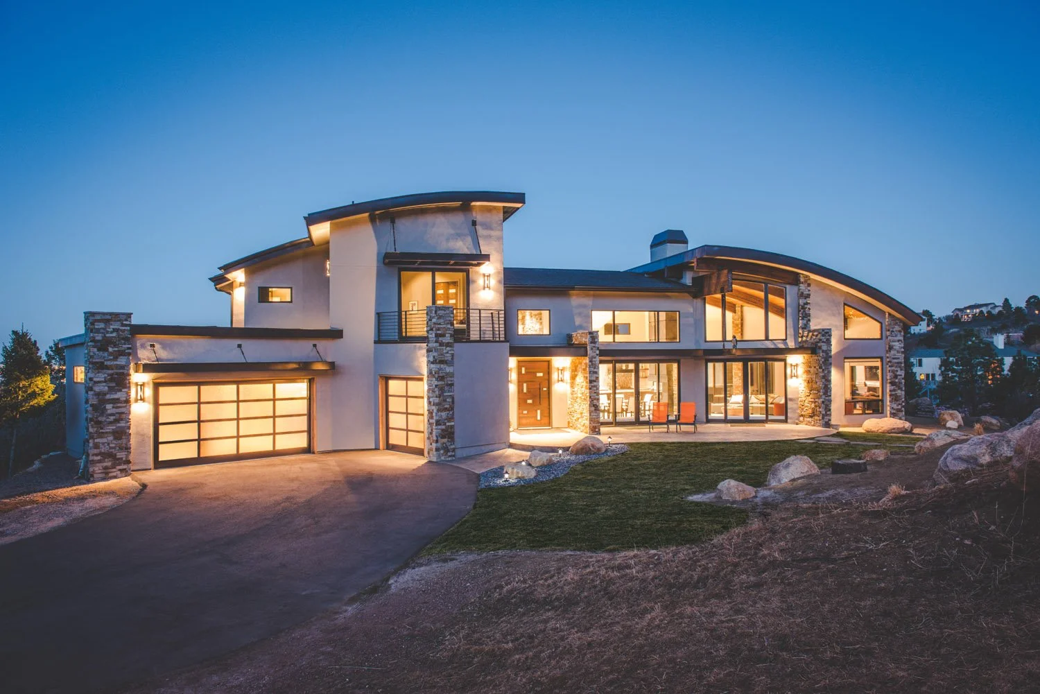 Modern luxury house with large windows and curved roof, illuminated at dusk. Custom home designed by LGA Studios in Broadmoor, Colorado Springs.