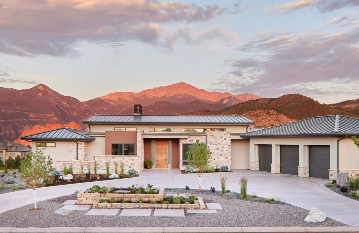 Modern house custom designed by LGA Studios with stone and glass facade, three-car garage, and Pikes Peak mountain backdrop at sunset in Kissing Camels, Colorado. 