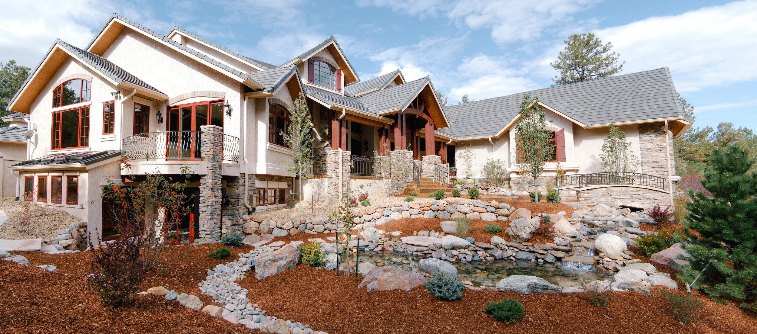 Large modern house with stone and stucco exterior, featuring numerous windows and a landscaped garden in the foreground, including a rock pathway and small pond.