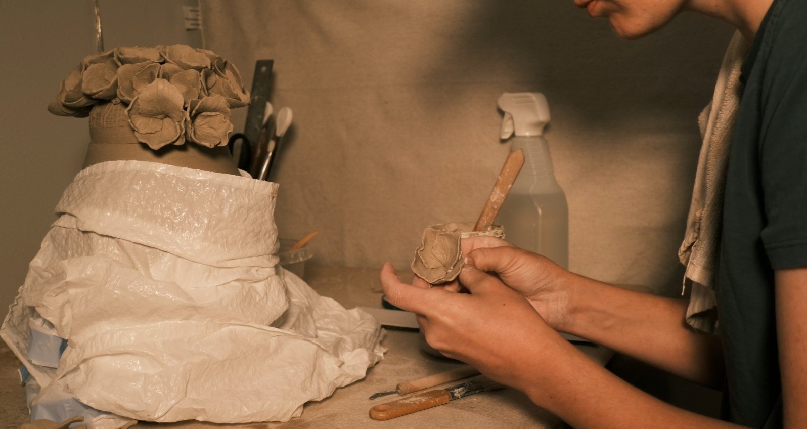 A person working on a sculpture of a hydrangea, shaping clay on a table inside a workshop.