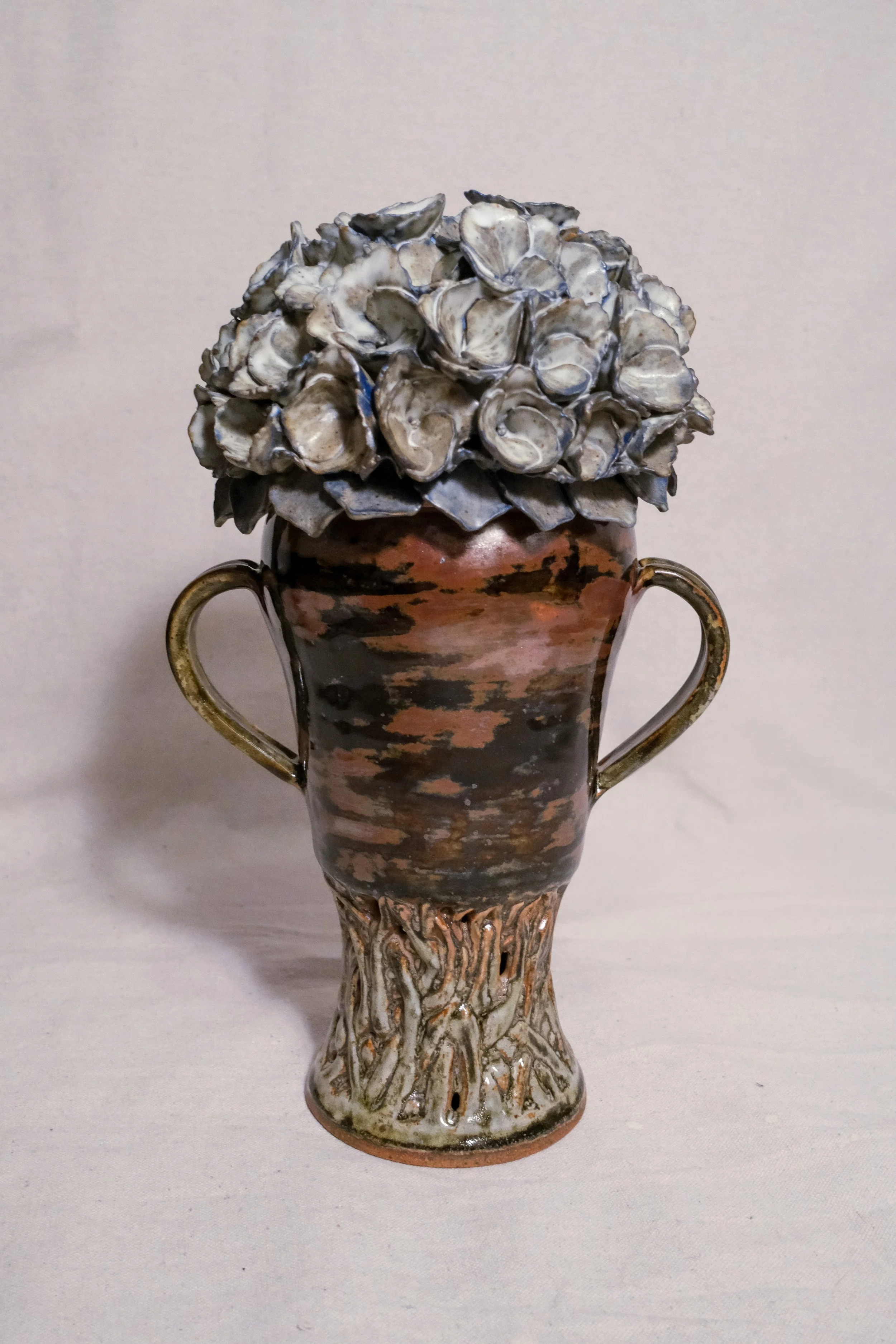 A ceramic vase with a black and brown pattern, featuring a handle on each side and a textured base resembling vines, the lid is a blue and white sculpted hydrangea