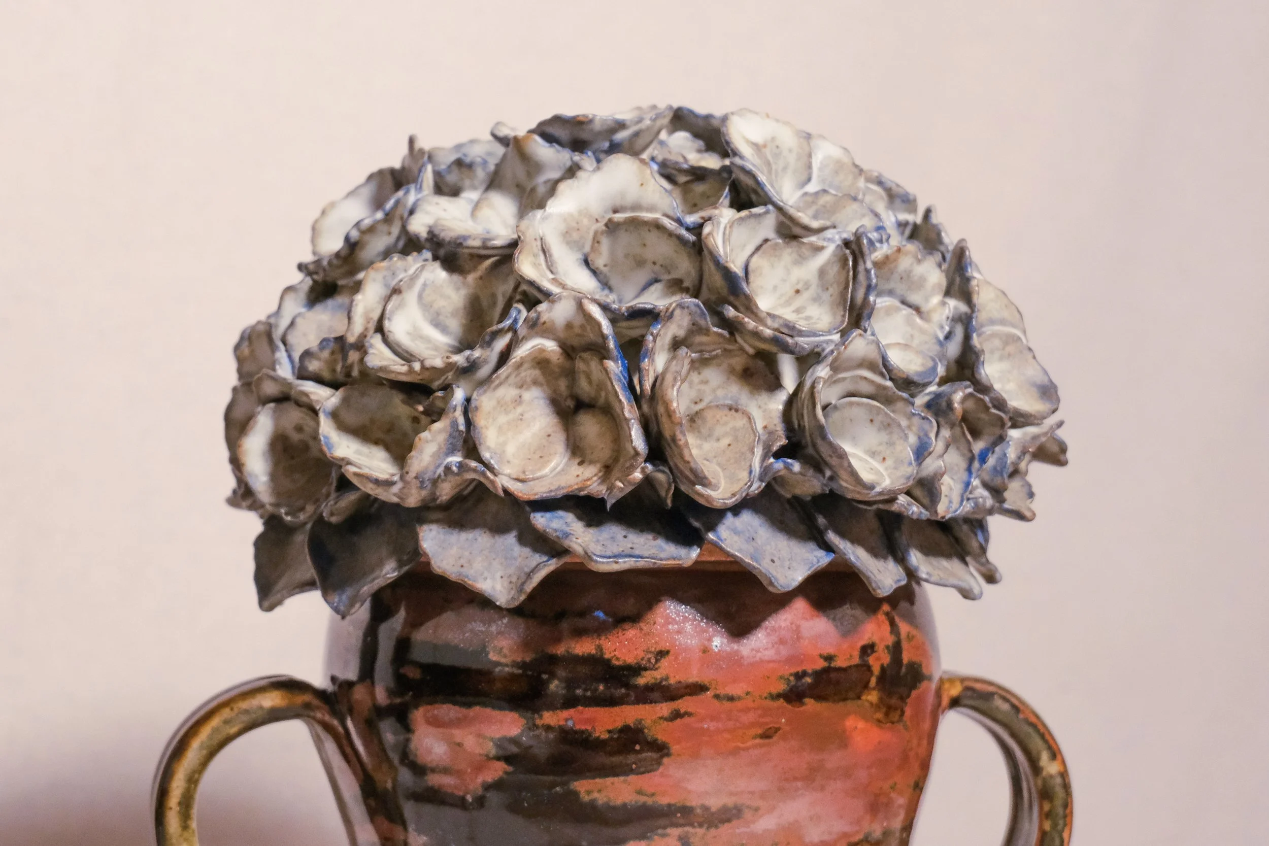 A close up of a ceramic vase with a black and brown pattern, featuring a handle on each side, and the lid is a blue and white sculpted hydrangea