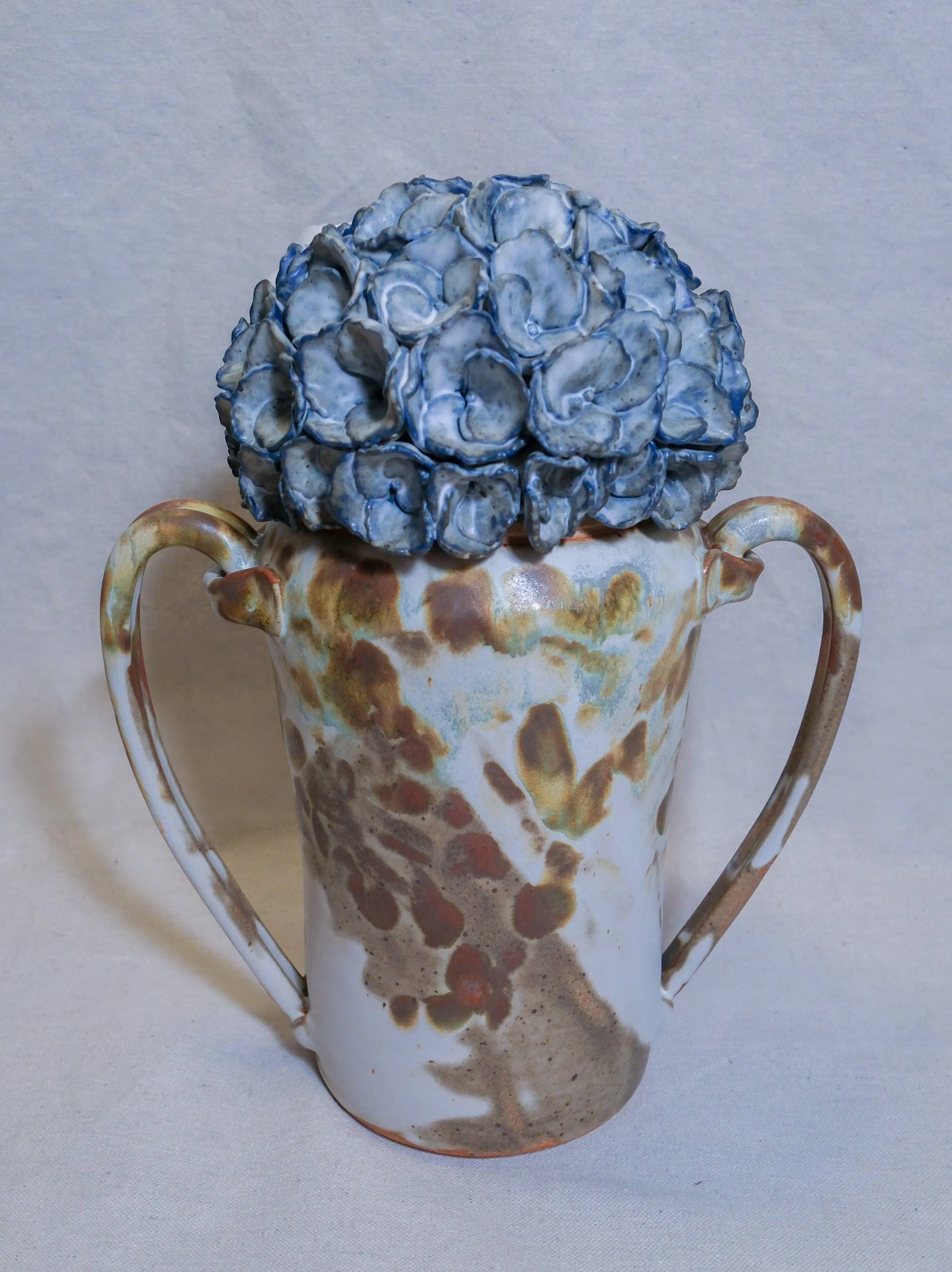 A decorative ceramic vase with a blue and white sculpted hydrangea lid, with a mottled brown and cream glaze. The vase has two curved handles on each side.