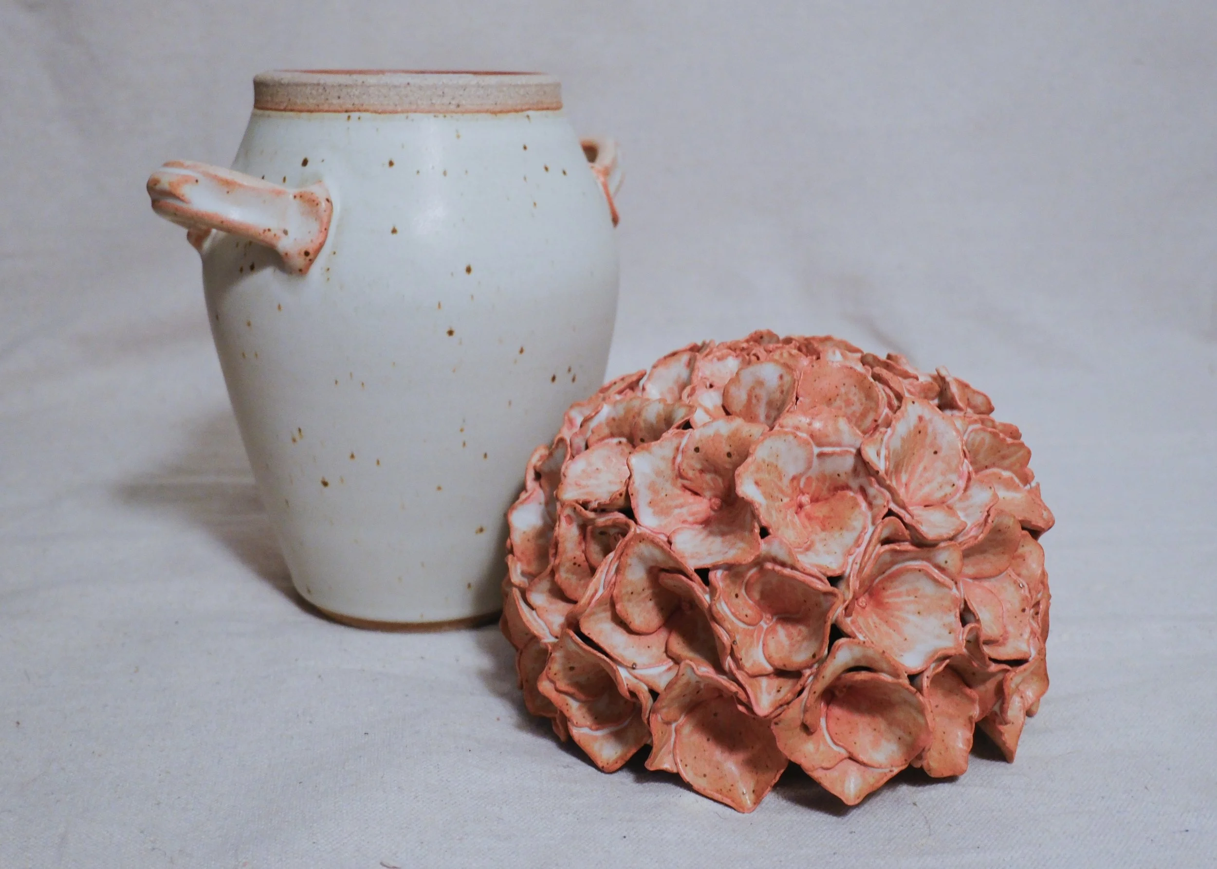 A white ceramic vase filled with a pink and white handle, and the lid is a dusty pink sculpted hydrangea where the lid is removed and sitting next to the vase