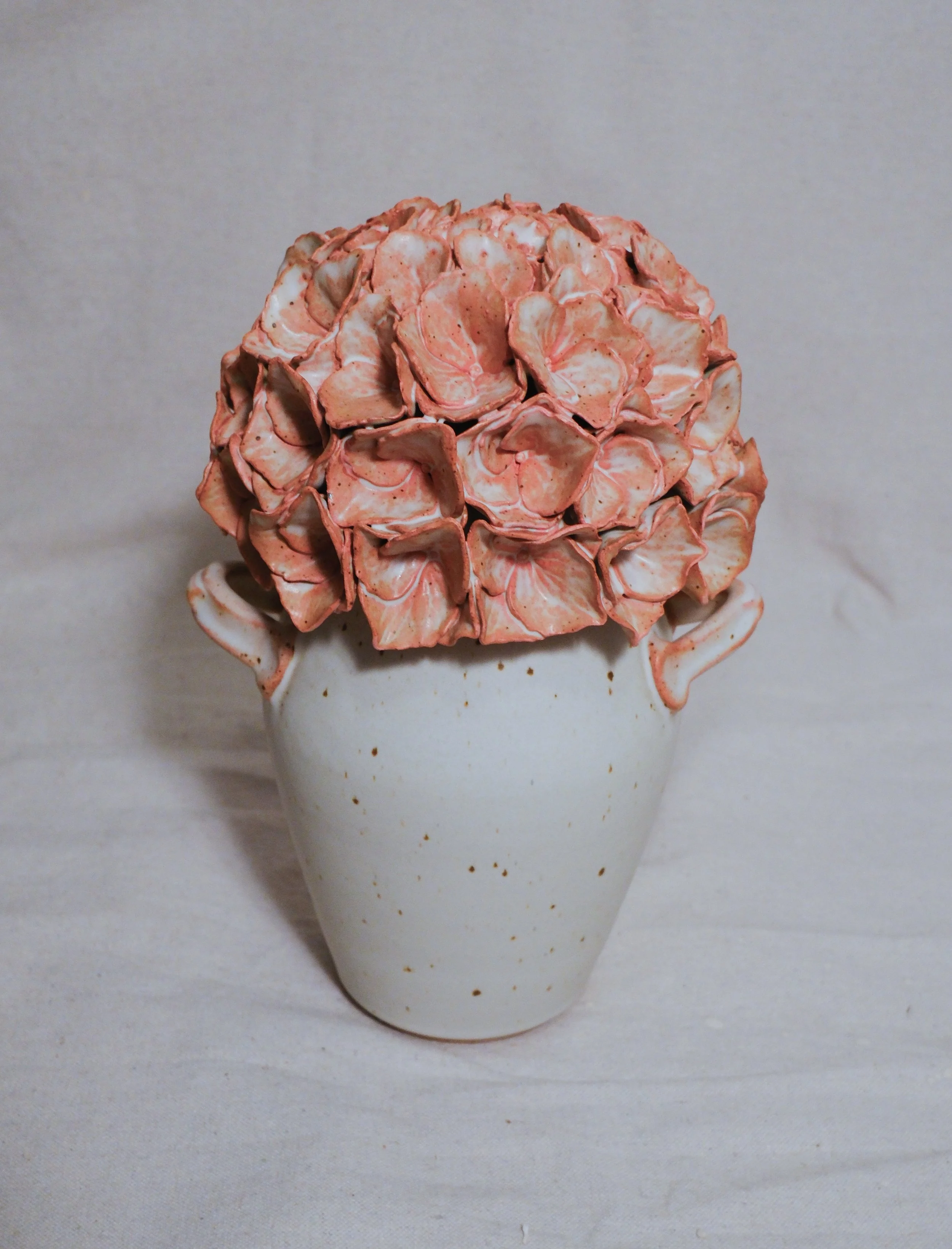 A white ceramic vase filled with a pink and white handle, and the lid is a dusty pink sculpted hydrangea, set against a plain background.