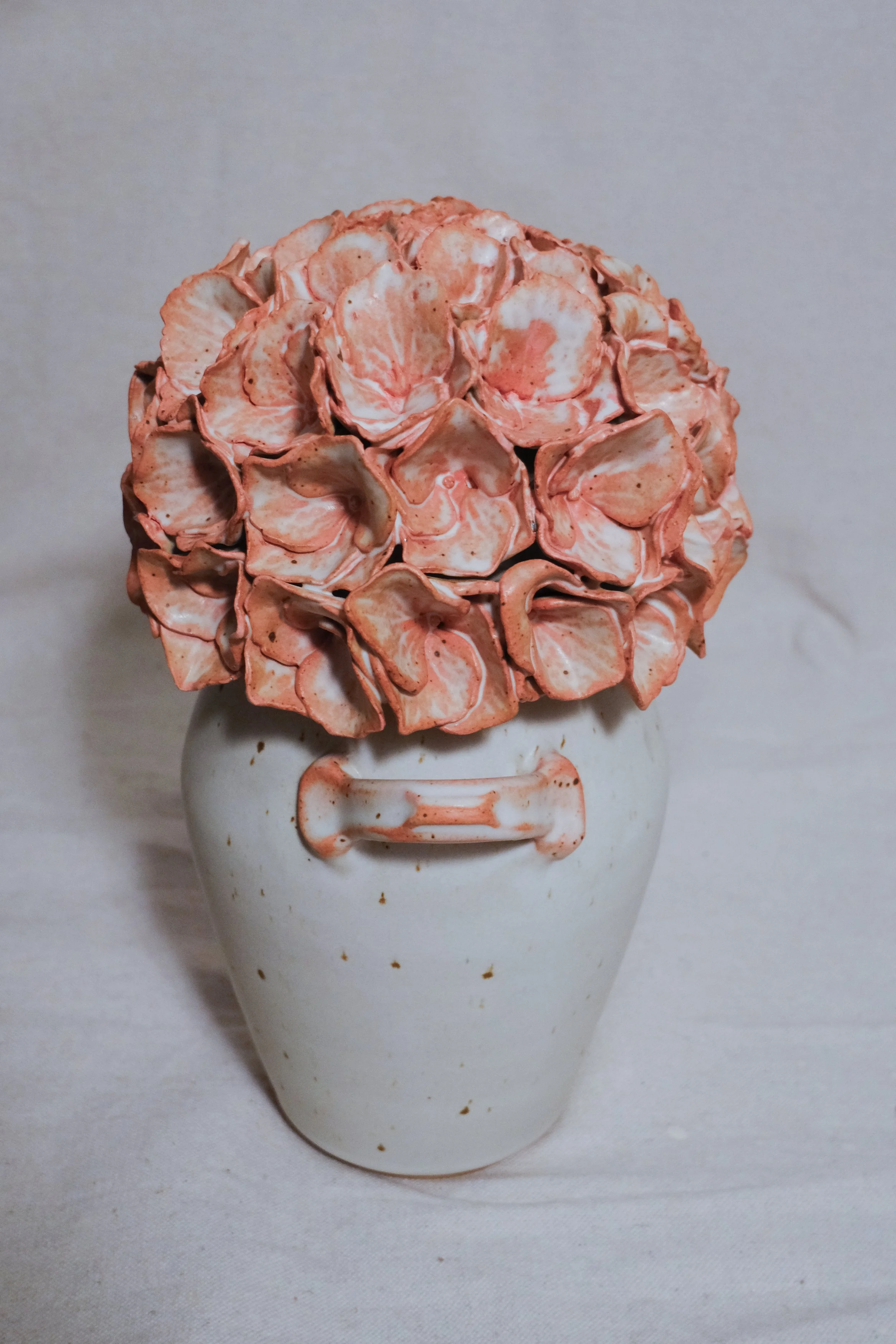 A white ceramic vase filled with a pink and white handle, and the lid is a dusty pink sculpted hydrangea, set against a plain background.