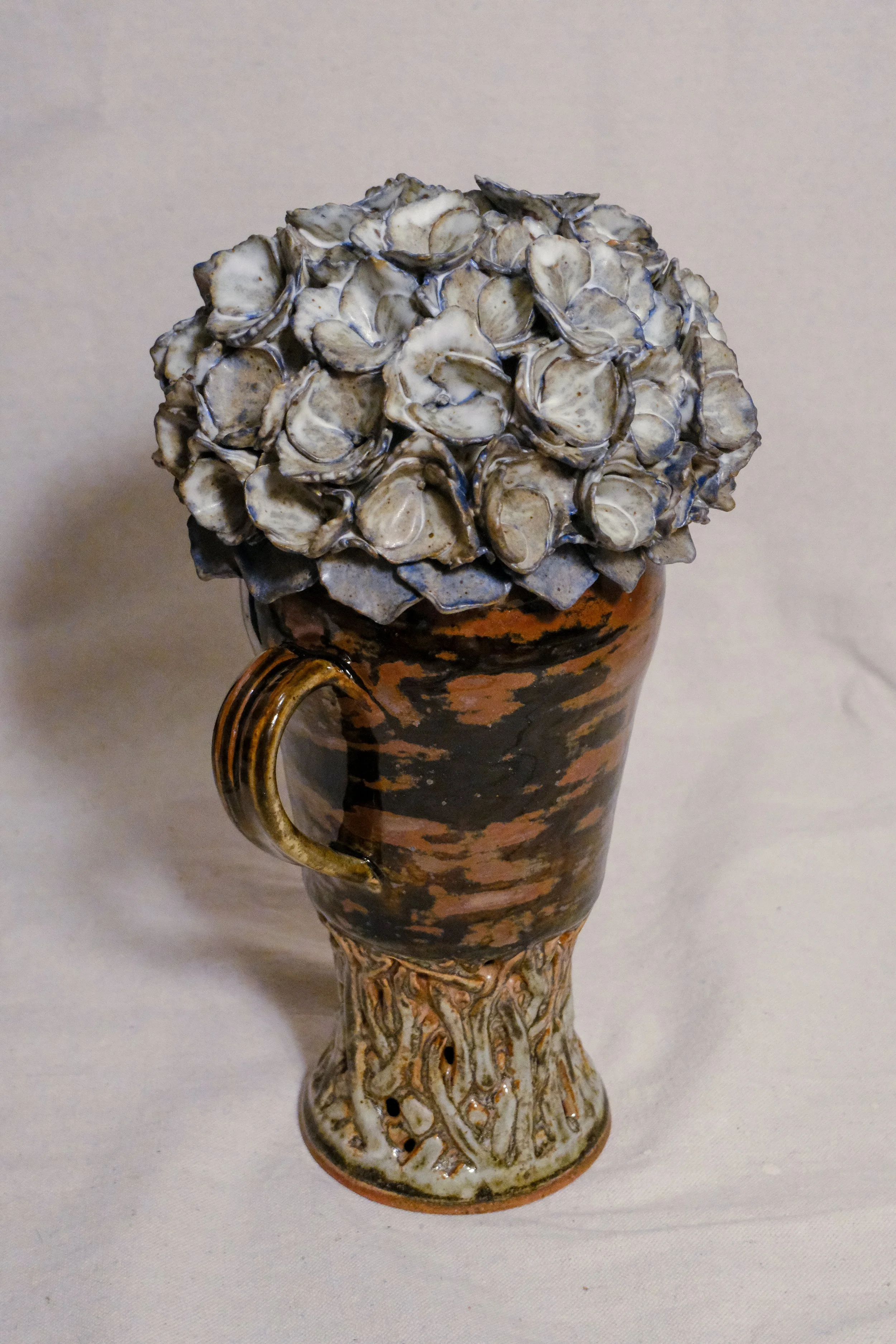 A ceramic vase with a black and brown pattern, featuring a handle on each side and a textured base resembling vines, the lid is a blue and white sculpted hydrangea