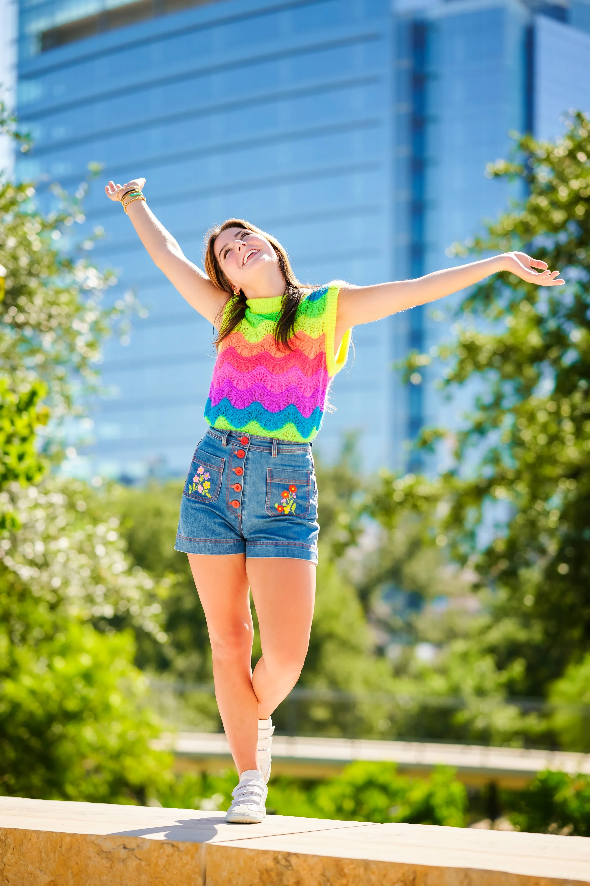 A young woman with arms outstretched, smiling, standing outdoors on a sunny day in front of greenery and modern blue buildings, wearing a colorful rainbow knitted sleeveless top, denim shorts with floral embroidery, and white sneakers.