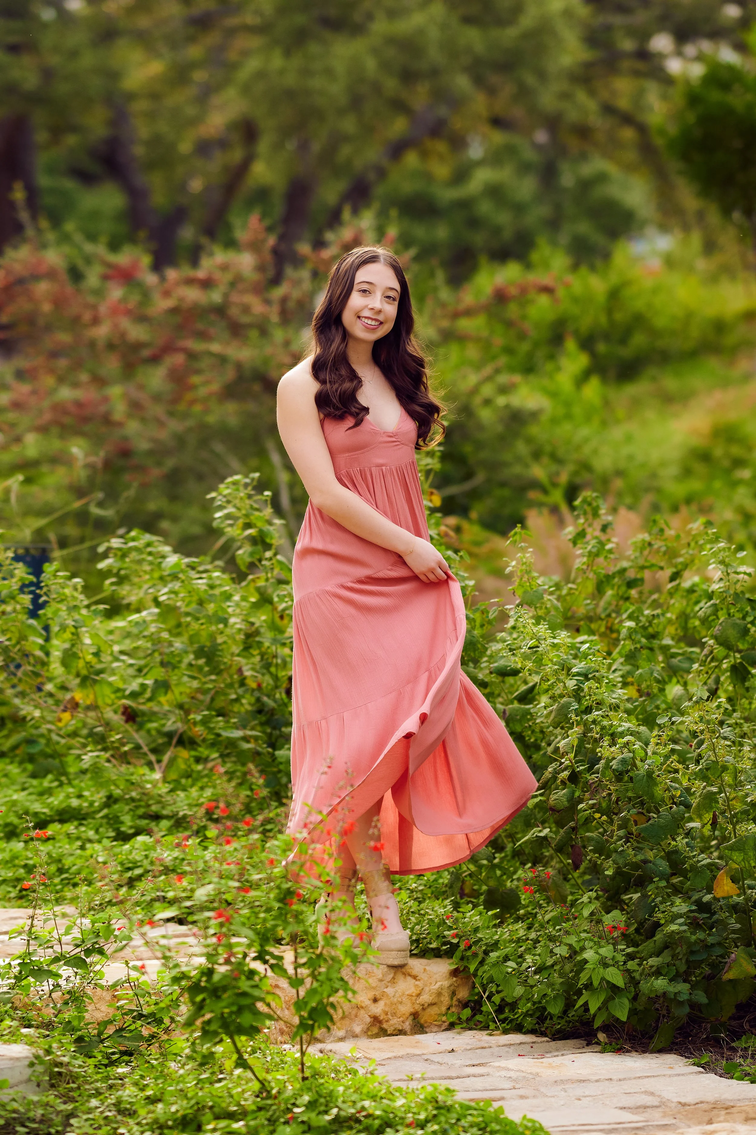 A young woman in a flowing pink dress standing on a stone pathway in a lush green garden or park.