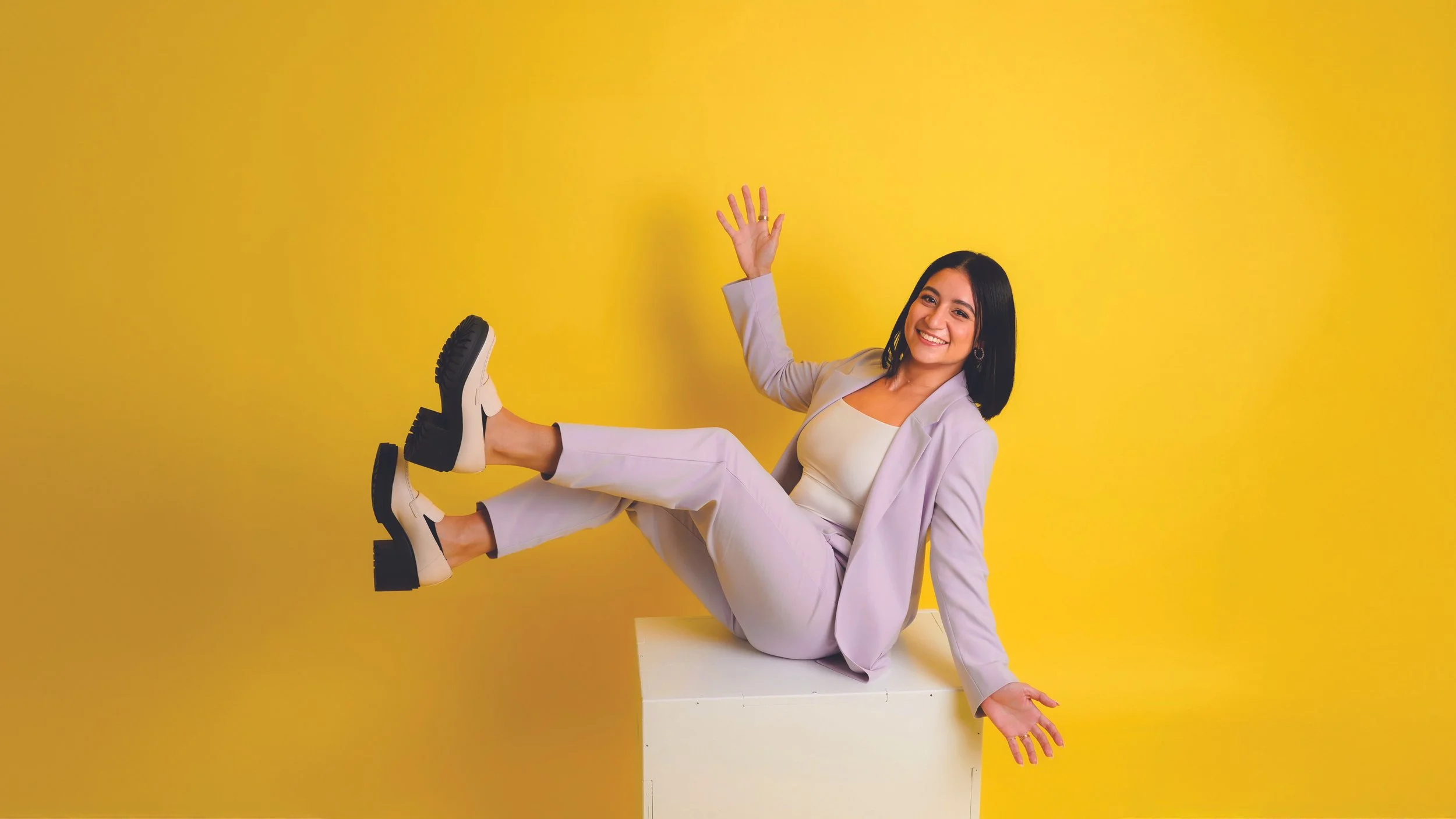 Smiling woman in a lavender suit sitting on a white block against a yellow background, waving with her right hand and holding her left hand out to the side.