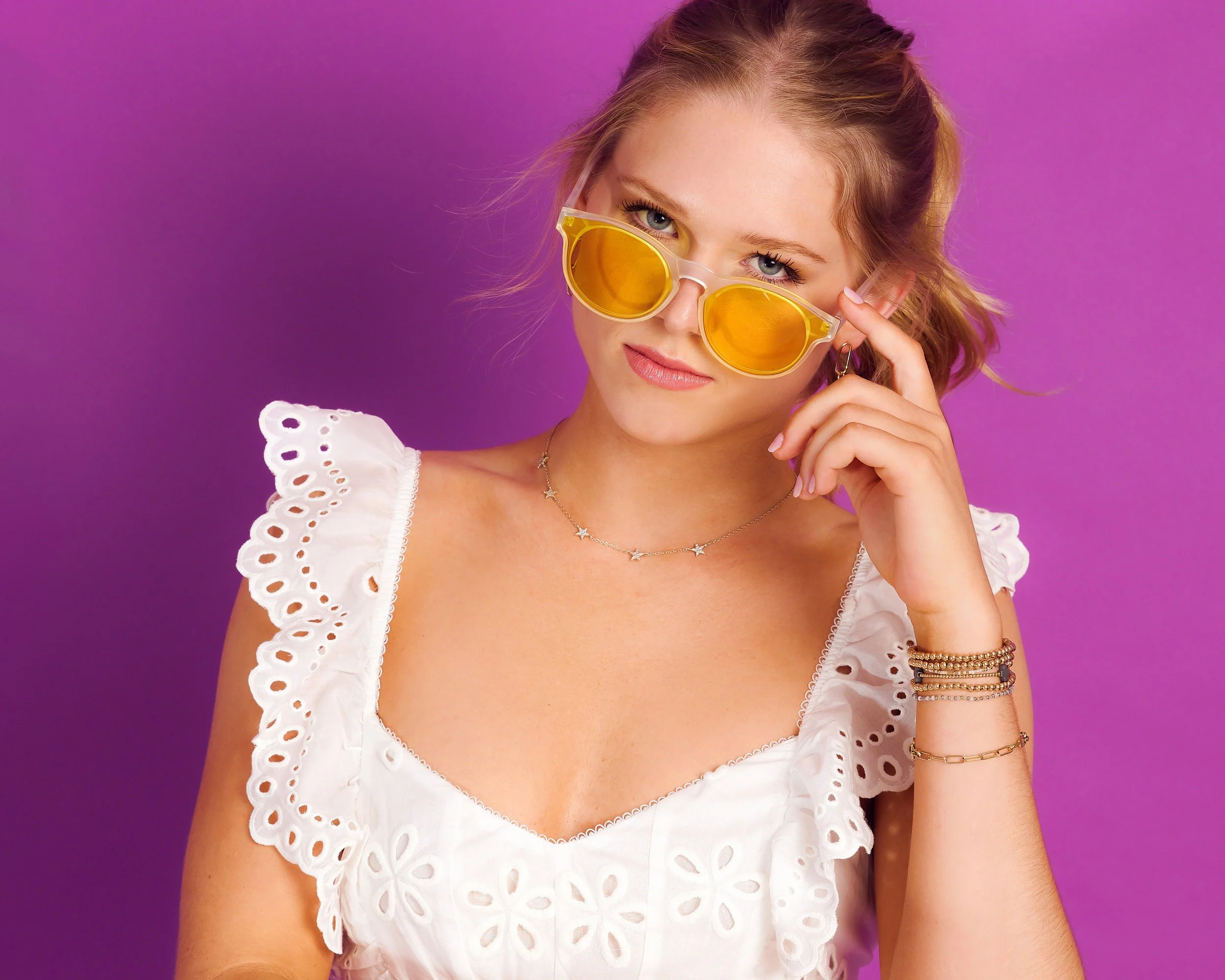 A young woman with wavy blonde hair, wearing yellow sunglasses, a white eyelet top, a star-shaped necklace, and multiple bracelets, posing against a purple background.